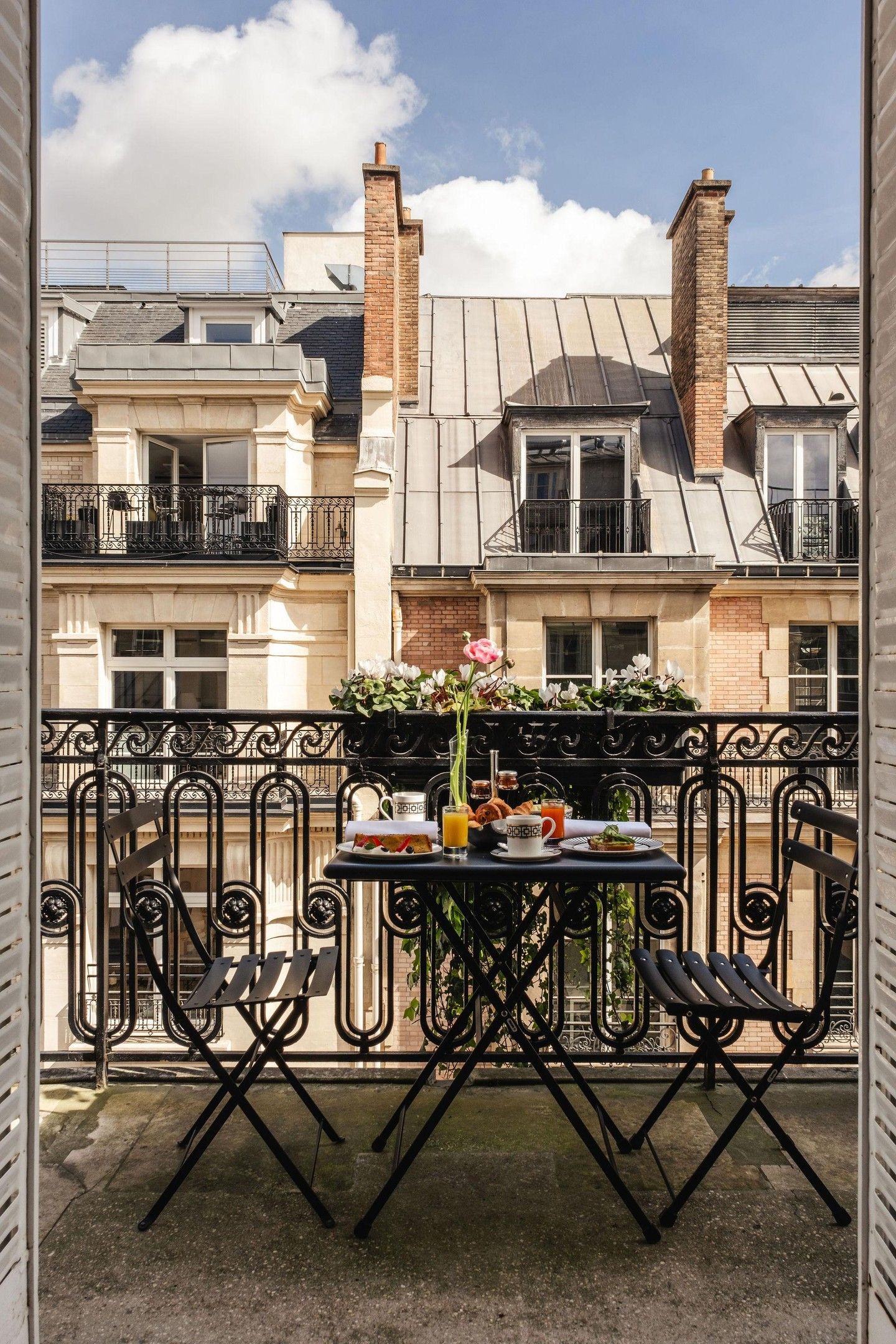 A balcony with a table set for breakfast, including a flower arrangement, a glass of orange juice, and a plate of food.
