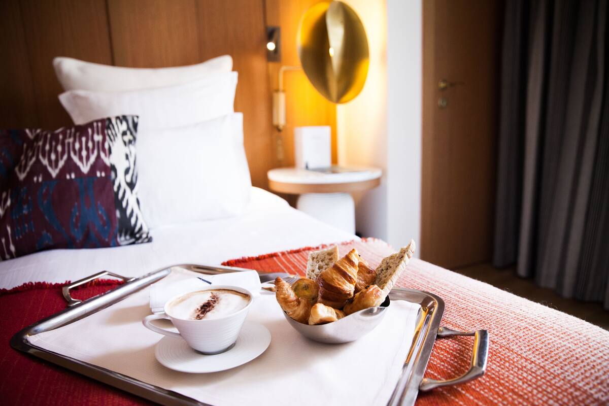 A hotel room with a bed, a bedside table with a lamp, a tray with croissants and coffee, and a pillow with a pattern.