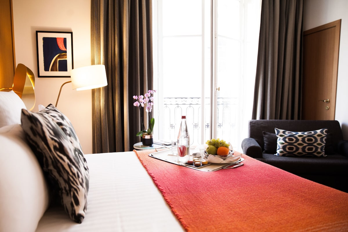 Ein Hotelzimmer mit einem Bett, einem Sofa, einem Tisch mit Obst und einer Flasche, einer Lampe, einem gerahmten Bild und Vorhängen.