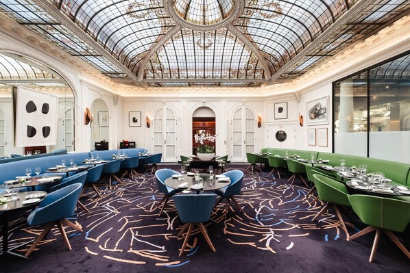 A restaurant with a high ceiling and ornate glass dome, featuring a central round table surrounded by blue and green upholstered booths. - Hotel Vernet (Paris)