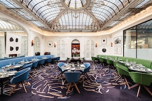 A restaurant with a high ceiling and ornate glass dome, featuring a central round table surrounded by blue and green upholstered booths. - Hotel Vernet (Paris)