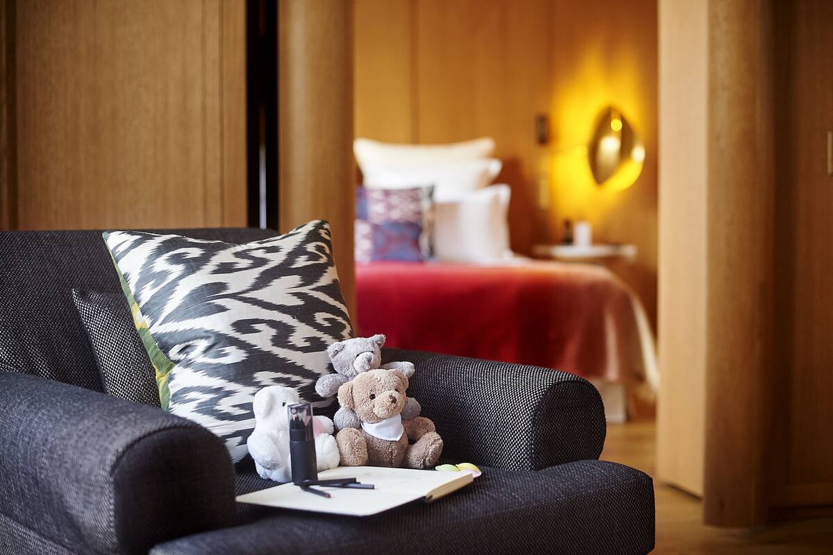 A room with a sofa, a bed with a red blanket, a nightstand with a lamp, and a teddy bear on the sofa.