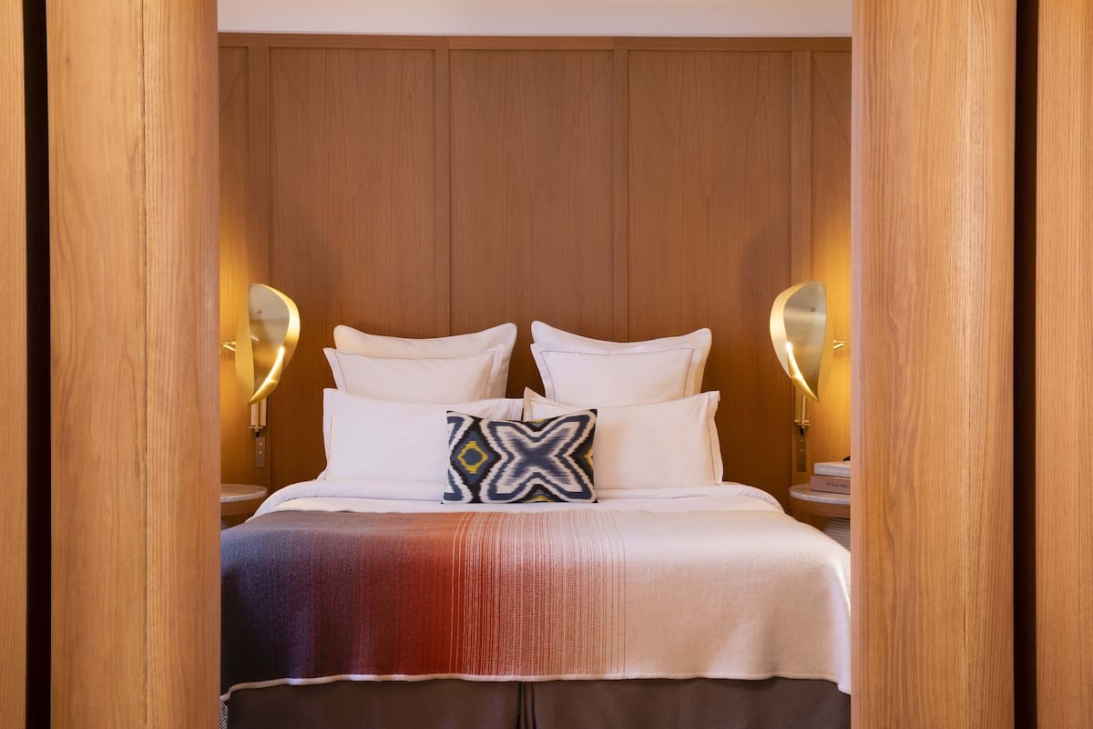 A neatly made bed with white linens and a patterned pillow, wooden headboard, and wall-mounted lamps.