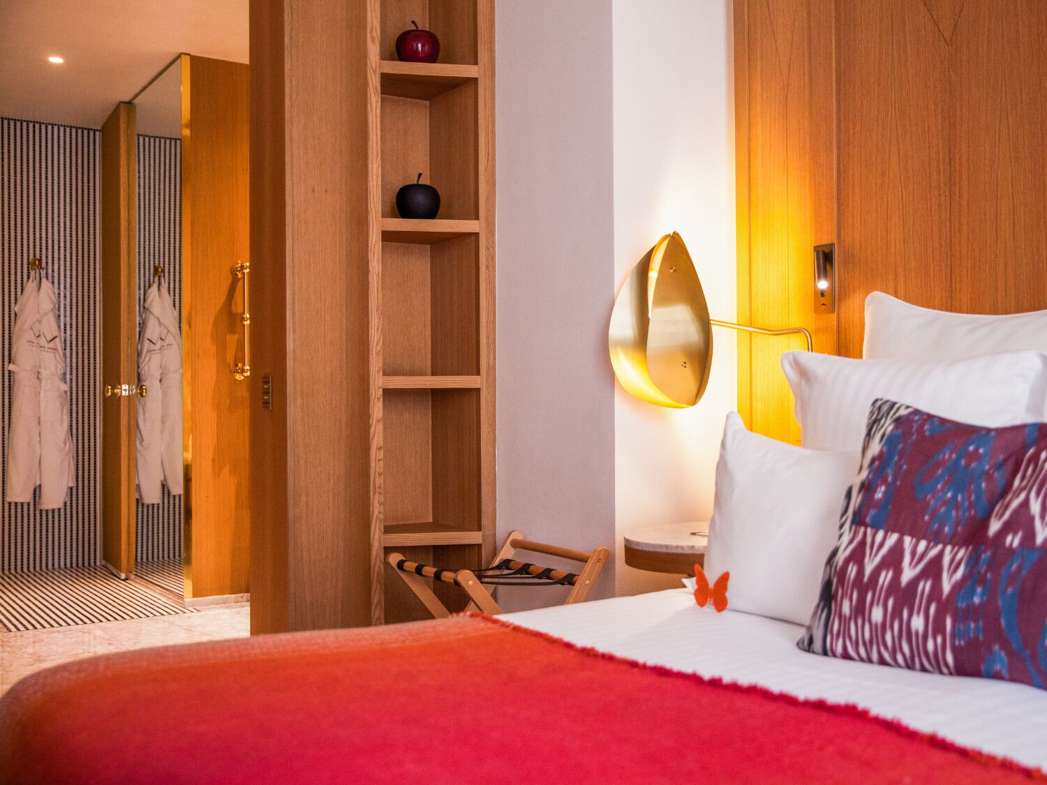 A hotel room with a bed, a wooden wardrobe, a mirror, a shelf with apples, a chair, and a robe rack.