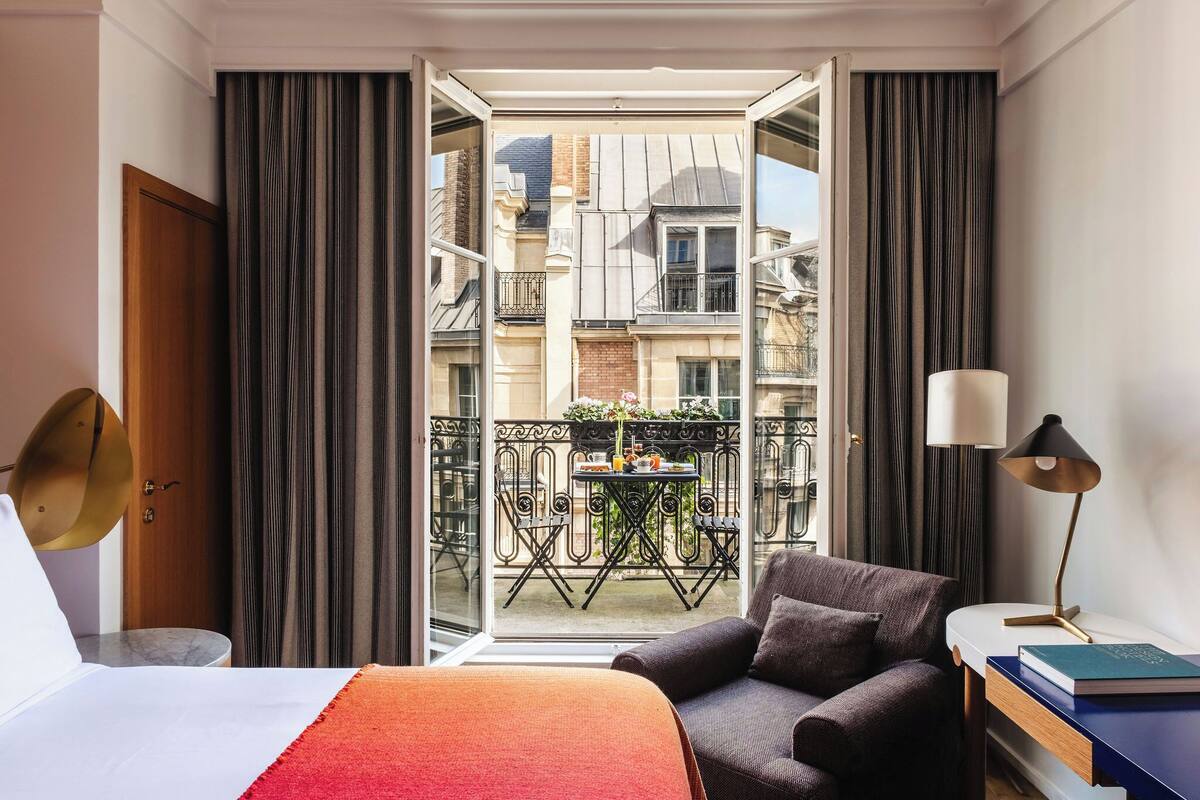 A hotel room with a bed, a chair, a table with a lamp, and a view of a balcony with a table and chairs outside.