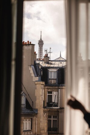 Vista a través de una ventana que muestra un edificio con una torre al fondo, un balcón y otra ventana con cortina.