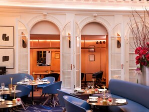 A hotel dining area with round tables, blue upholstered seating, and a view into the kitchen. - Hotel Vernet (Paris)