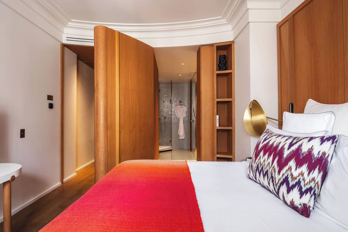 A hotel room with a bed, a bedside table, a wardrobe, and a bathroom visible through an open door.