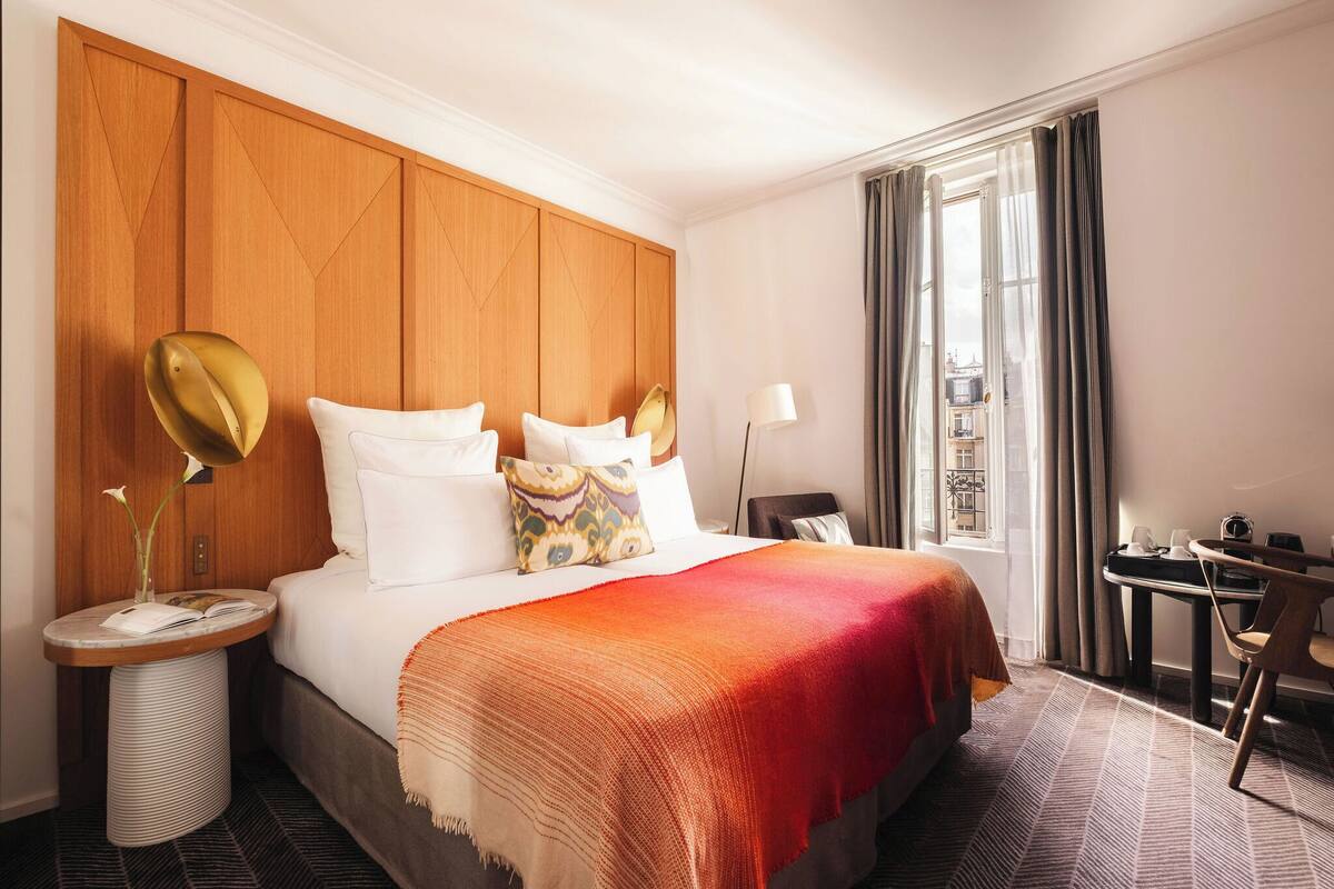 A hotel room with a bed, bedside table, lamp, and a view through the window.