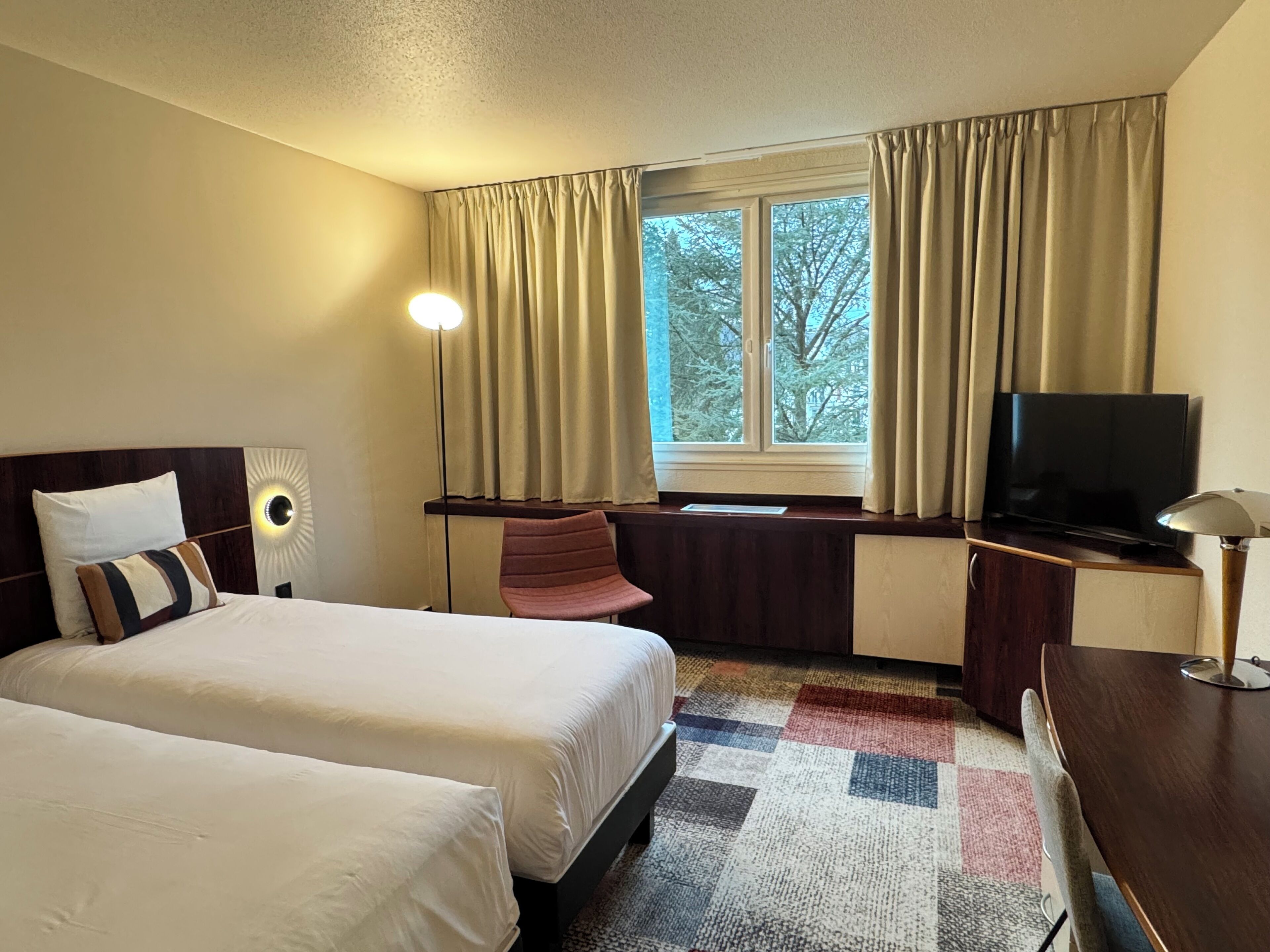 A hotel room with two beds, a desk, a TV, and a window with curtains.