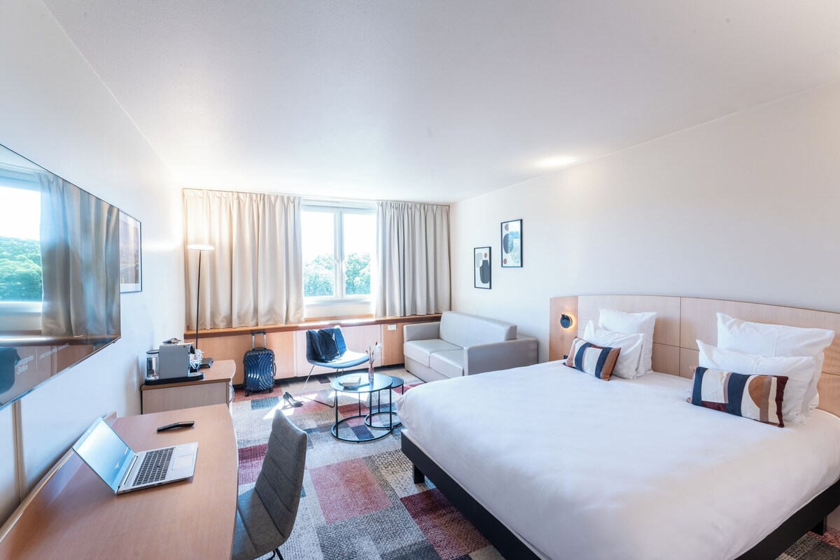 Una habitación de hotel moderna con una cama grande, un escritorio con una computadora portátil, una silla, una mesita redonda y un ventanal con cortinas.