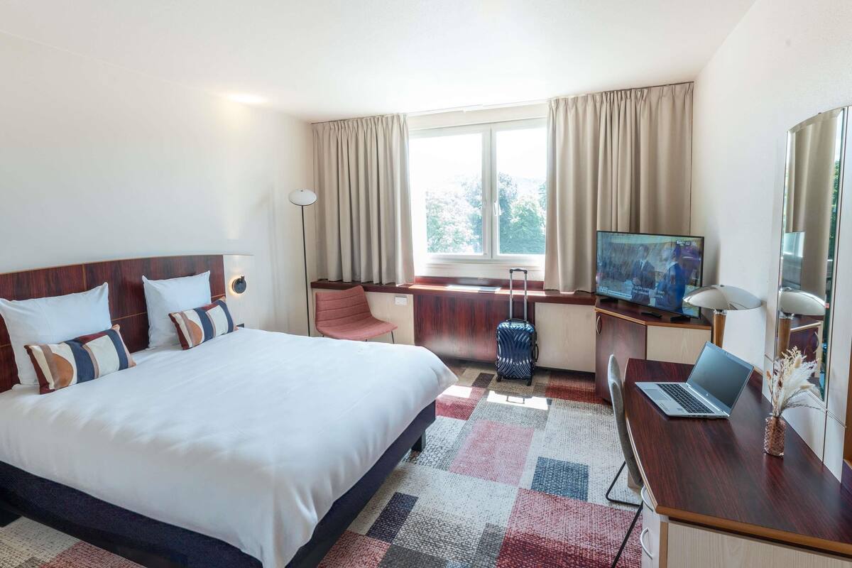 A hotel room with a bed, a desk with a laptop, a television, and a suitcase.