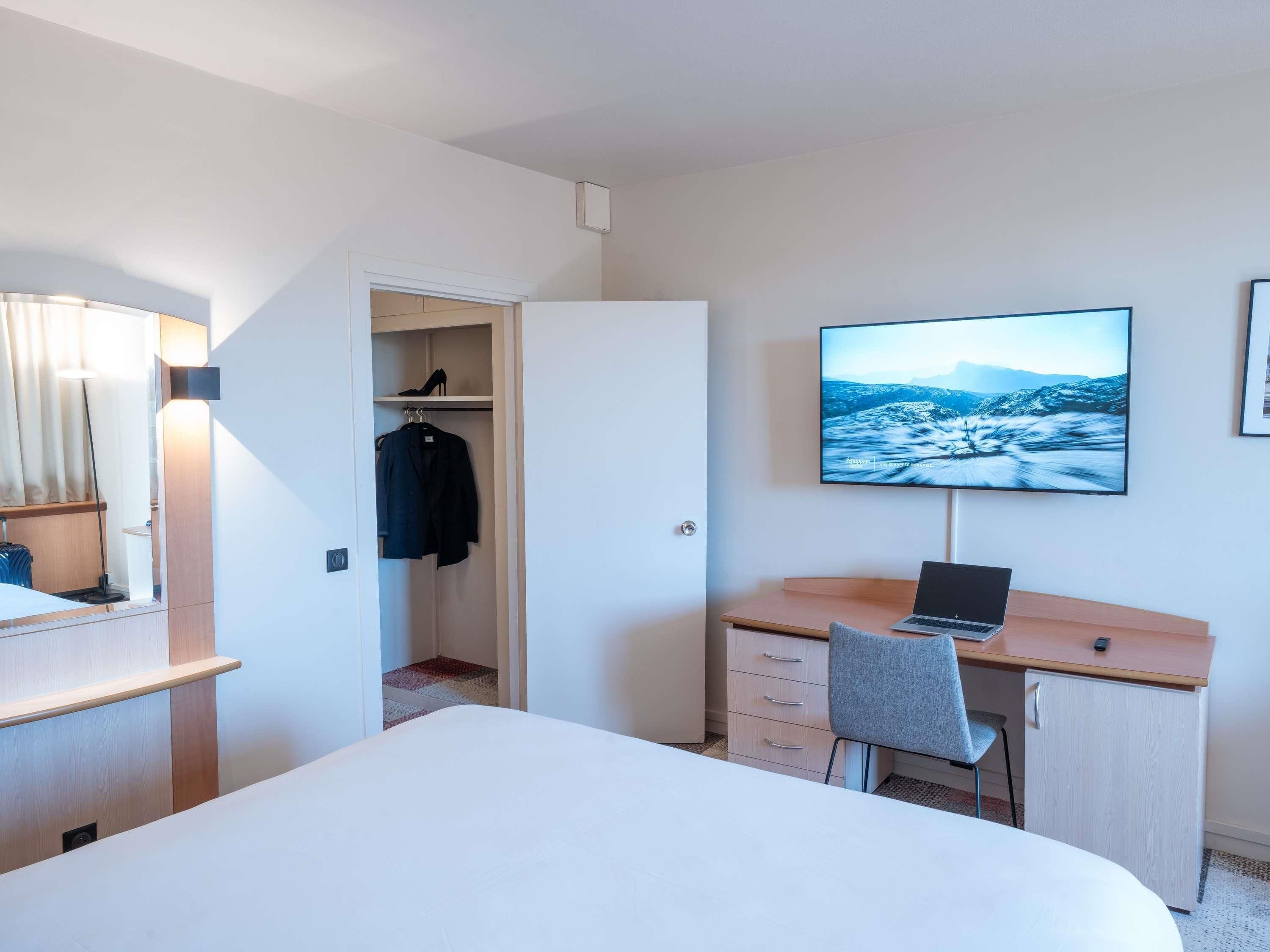 A hotel room with a bed, a desk with a laptop, a wardrobe, and a TV on the wall.