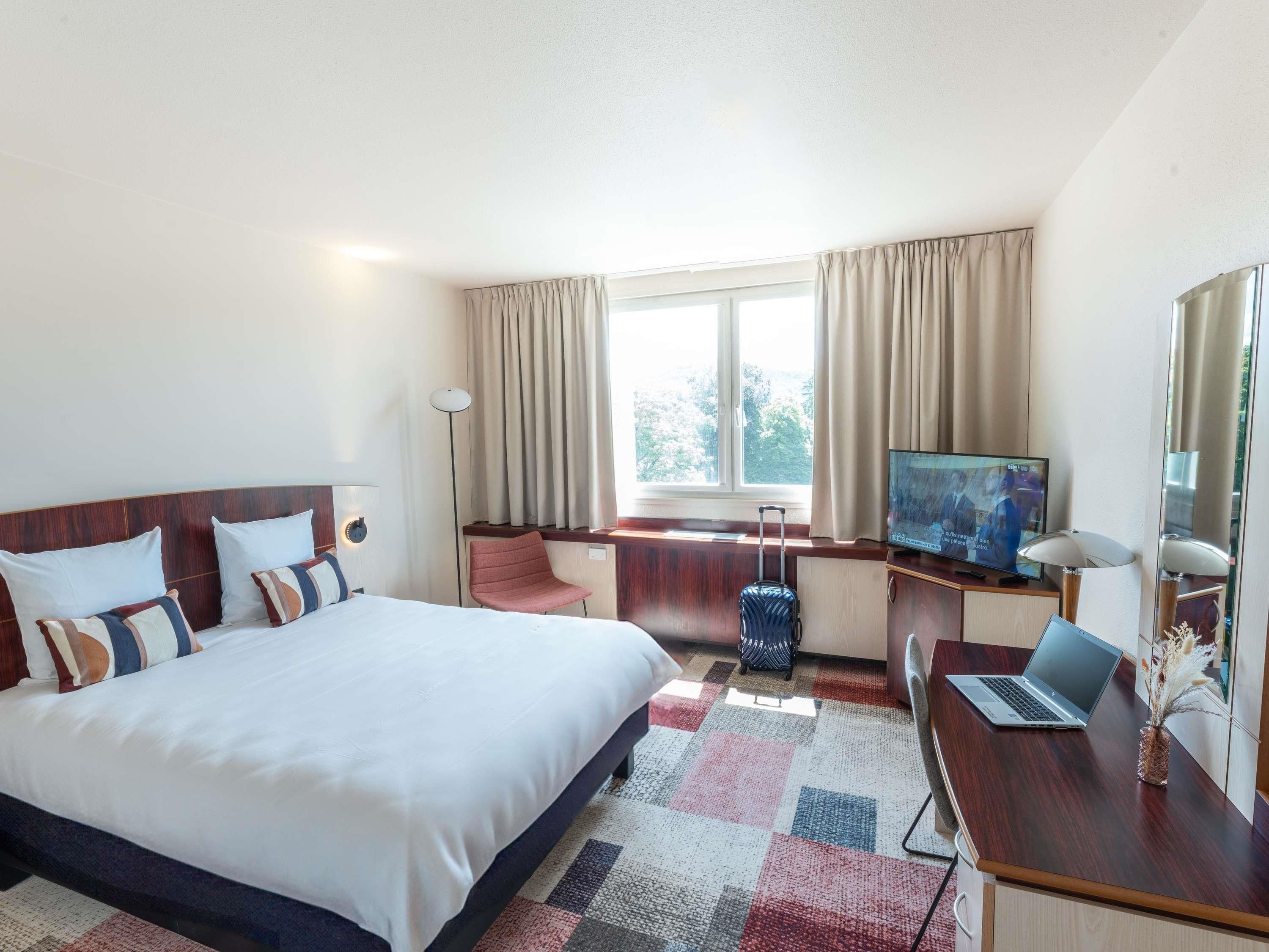 A hotel room with a bed, a desk with a laptop, a television, and a suitcase.