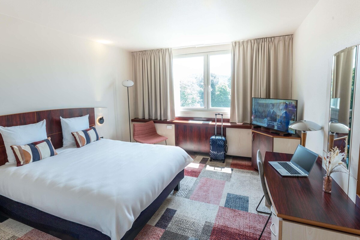 Una habitación de hotel con cama, escritorio, computadora portátil, televisor y maleta.