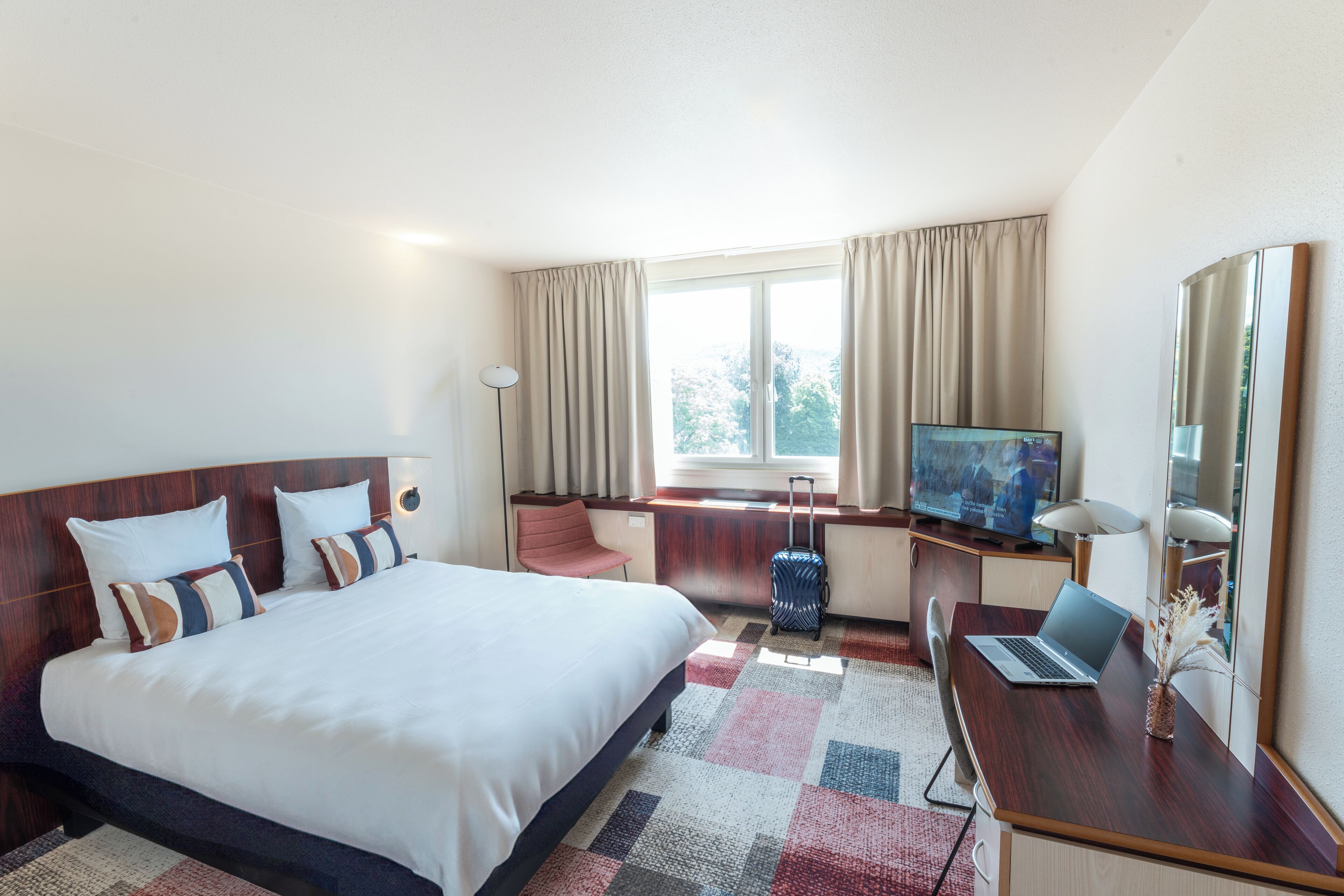 Una habitación de hotel con una cama grande, un escritorio con una computadora portátil, un televisor y una maleta.