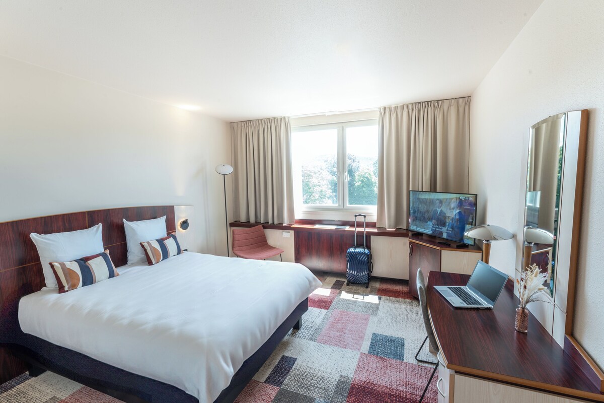 Una habitación de hotel con una cama grande, un escritorio con una computadora portátil, un televisor y una maleta.