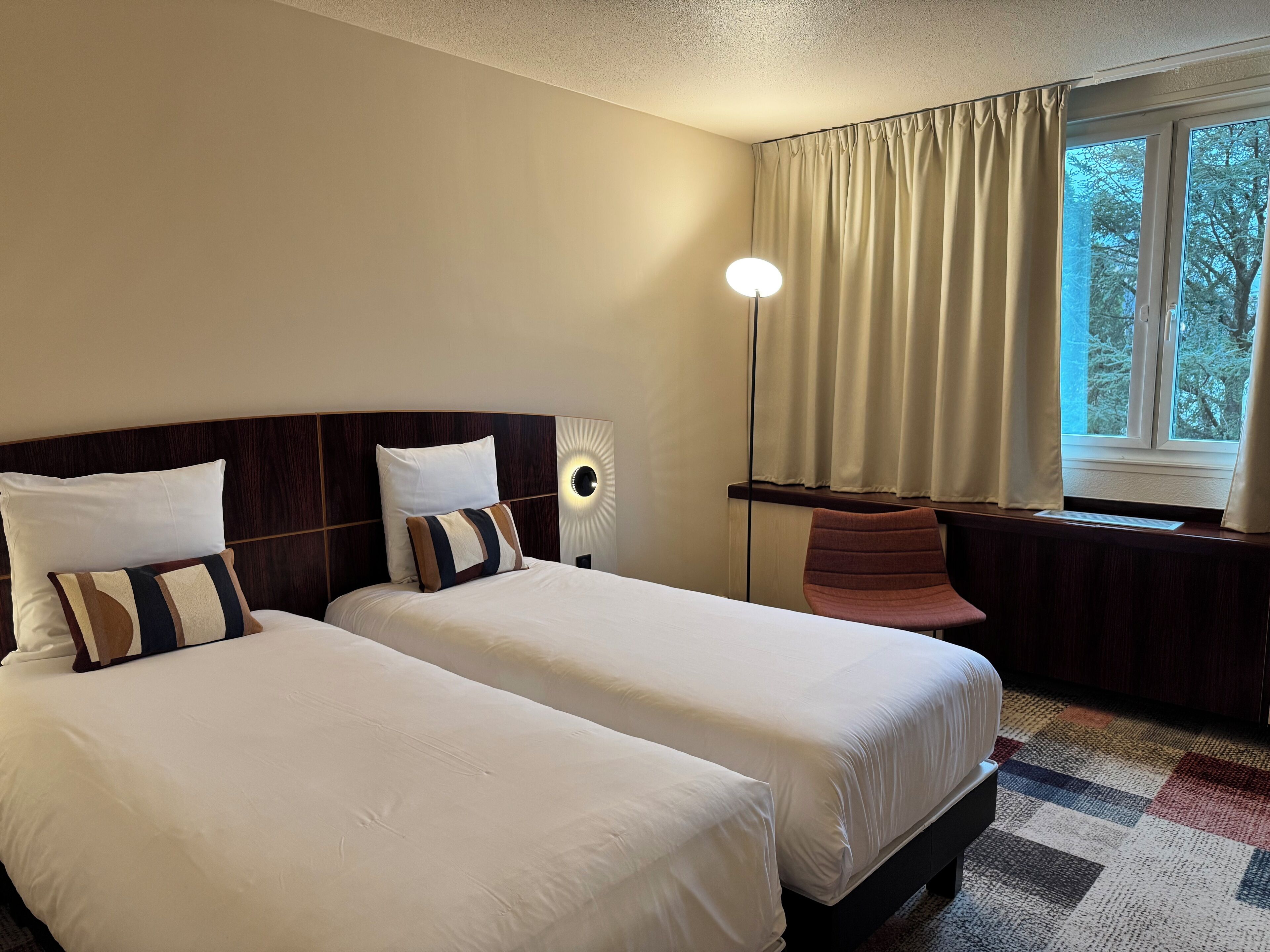 A hotel room with two beds, a nightstand, a lamp, a chair, and a window with curtains.