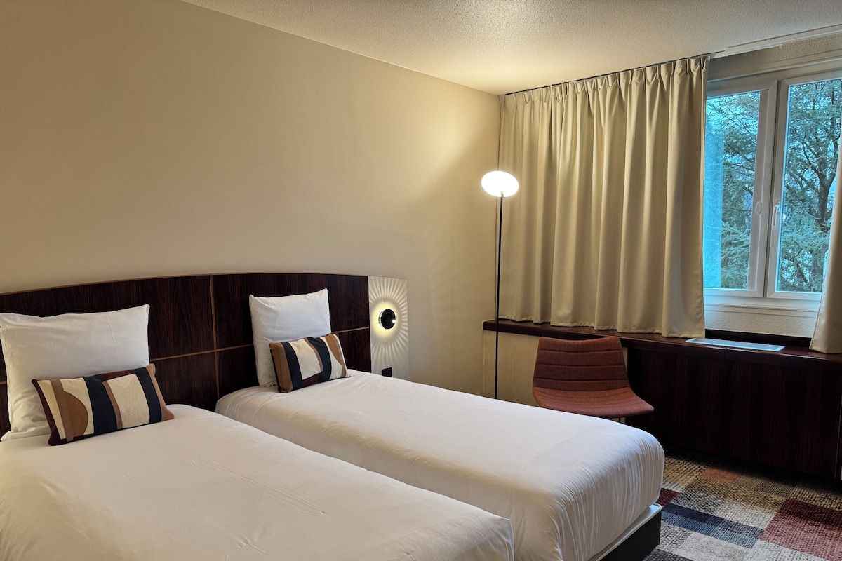 Habitación de hotel con dos camas, una mesita de noche, una lámpara, una silla y una ventana con cortinas.