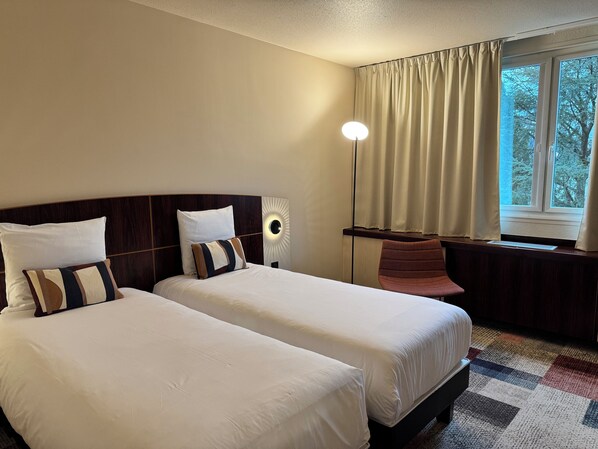 A hotel room with two beds, a nightstand, a lamp, a chair, and a window with curtains.