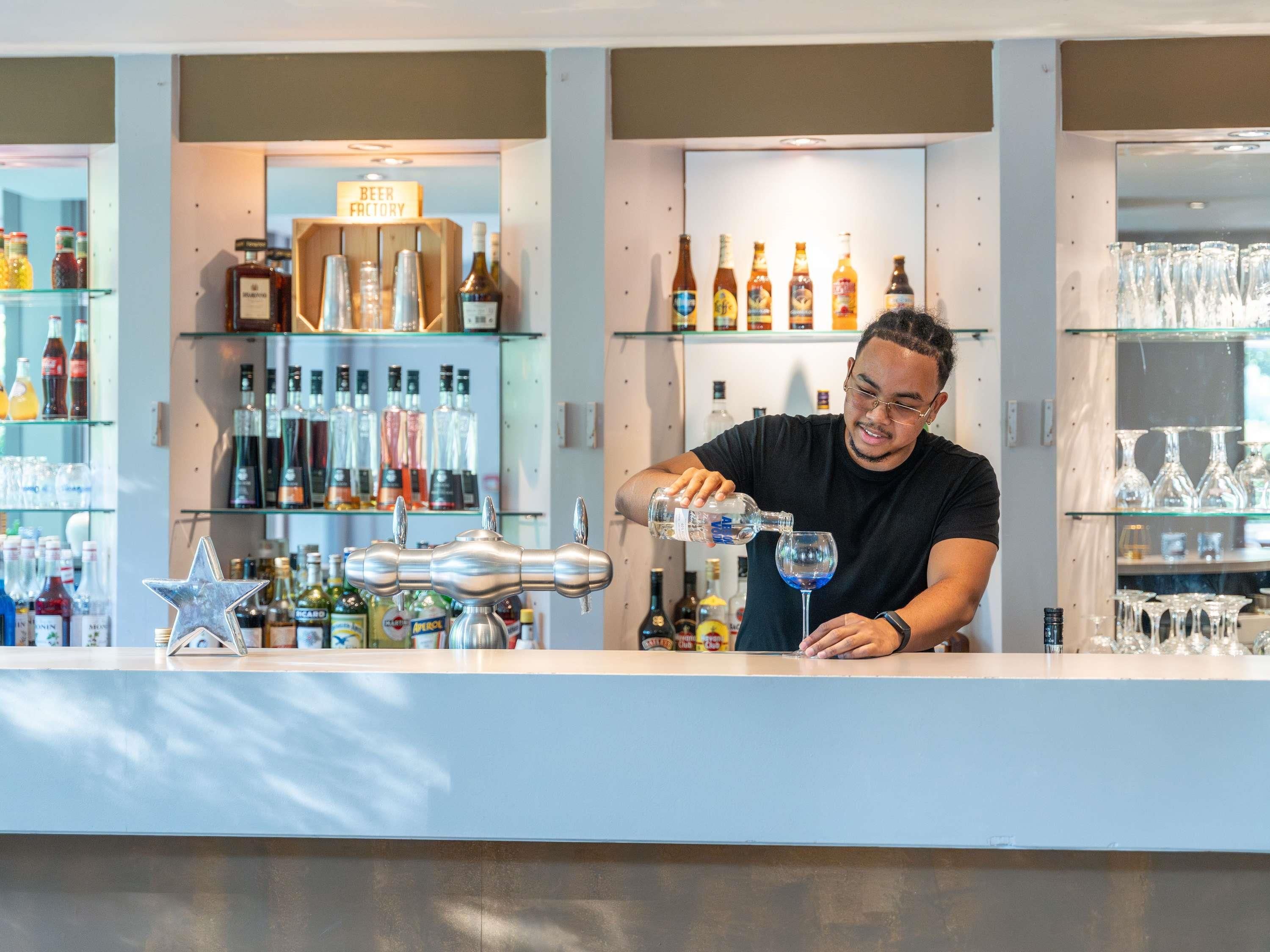 A bar area with a bartender pouring a drink into a glass. There are shelves with various bottles and glasses behind the bartender.