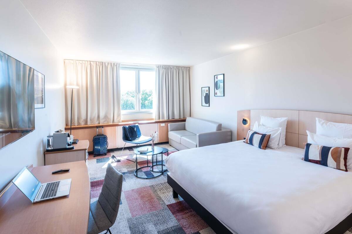 A hotel room with a bed, a desk with a laptop, a chair, a sofa, a small table, and a window with curtains.