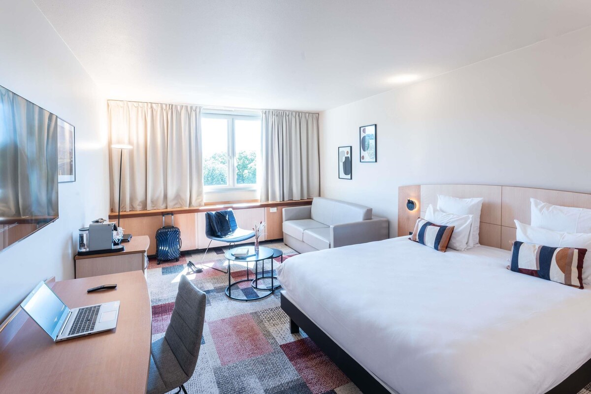 Habitación de hotel con cama, escritorio con una computadora portátil, silla, sofá, una mesita y ventana con cortinas.