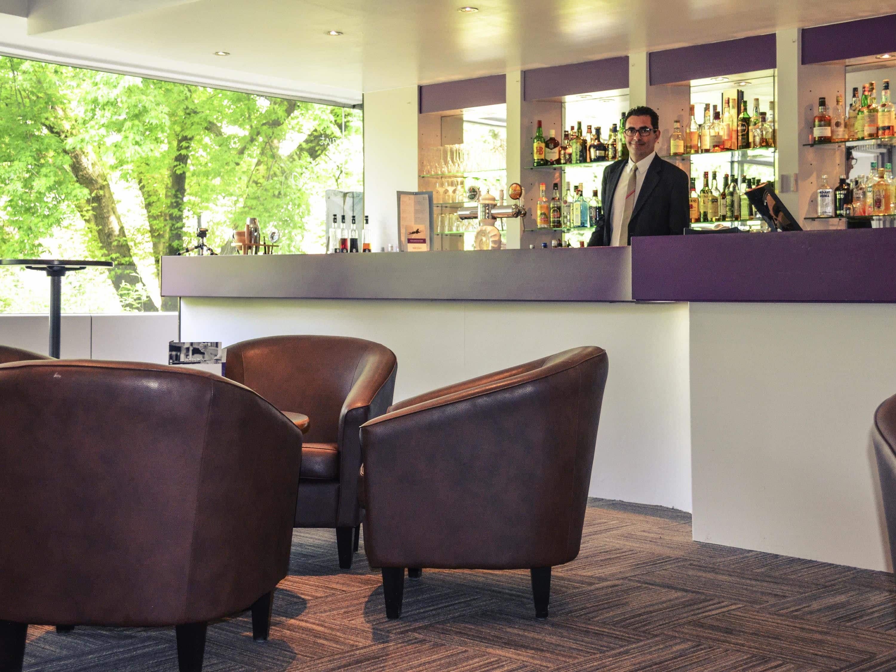A bar area with leather chairs, a counter with various bottles, and a view of trees through large windows.