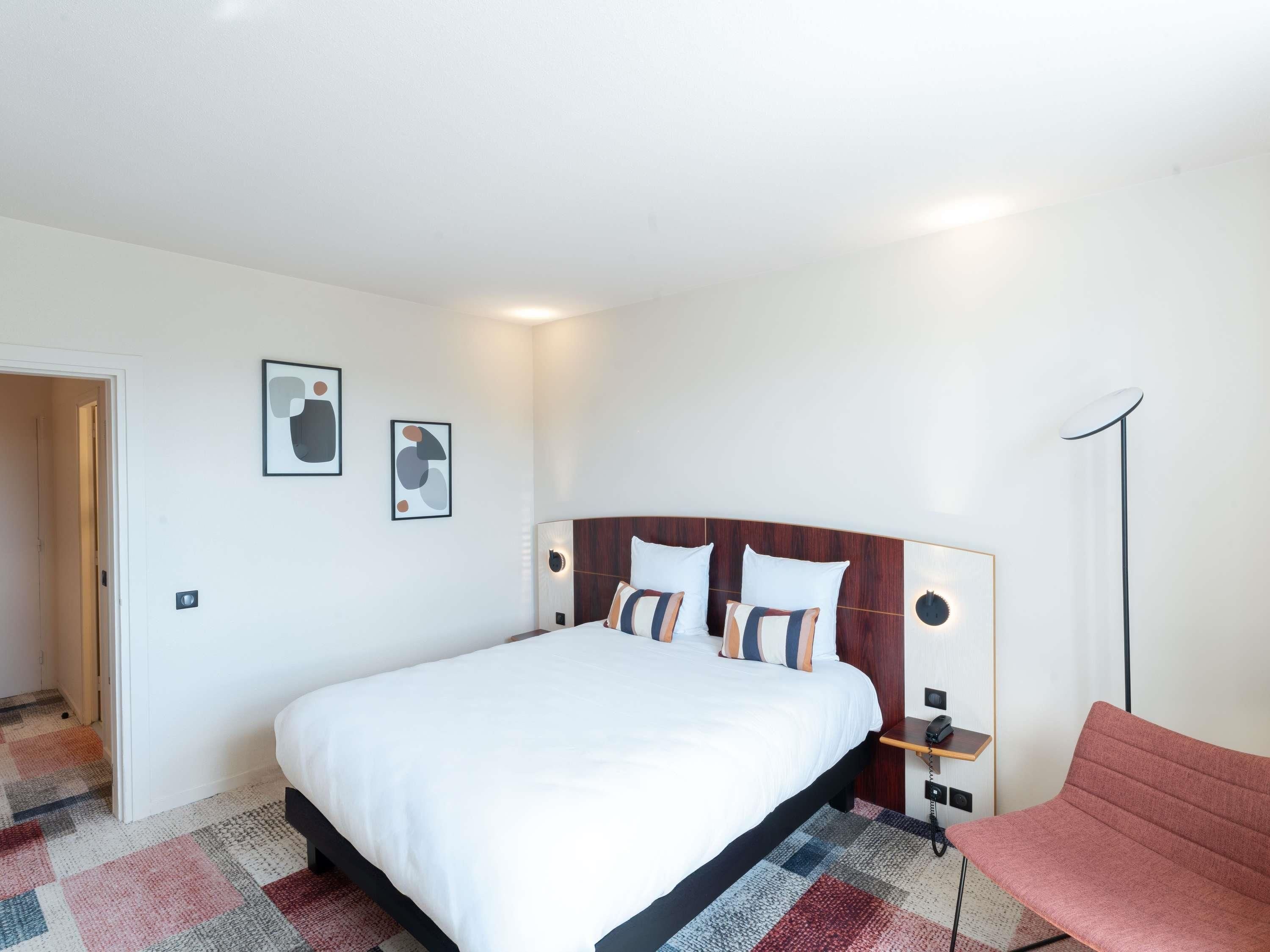 A hotel room with a large bed, a bedside table with a lamp, a chair, and abstract wall art.