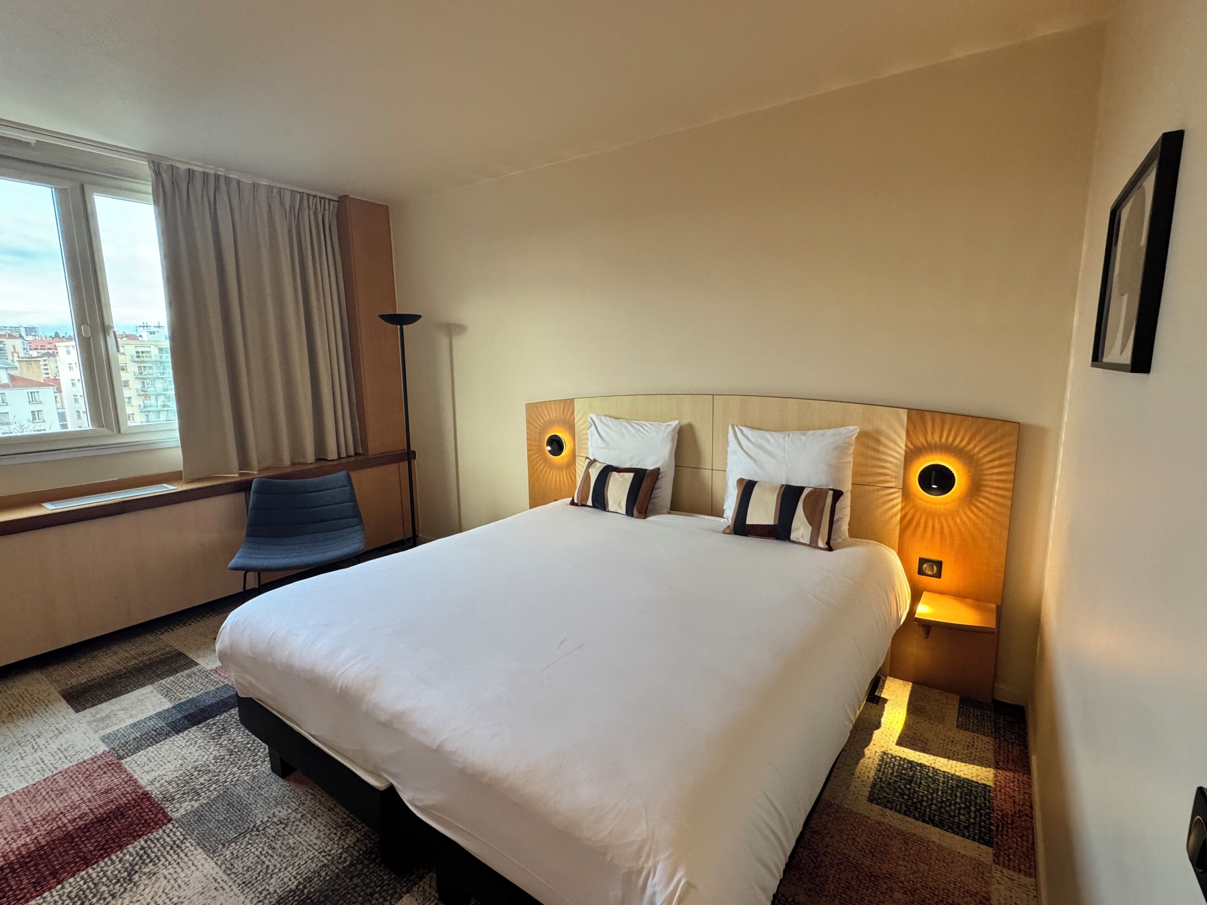 Habitación de hotel con una cama grande, dos lámparas de noche, una silla y vistas de la ciudad a través de la ventana.