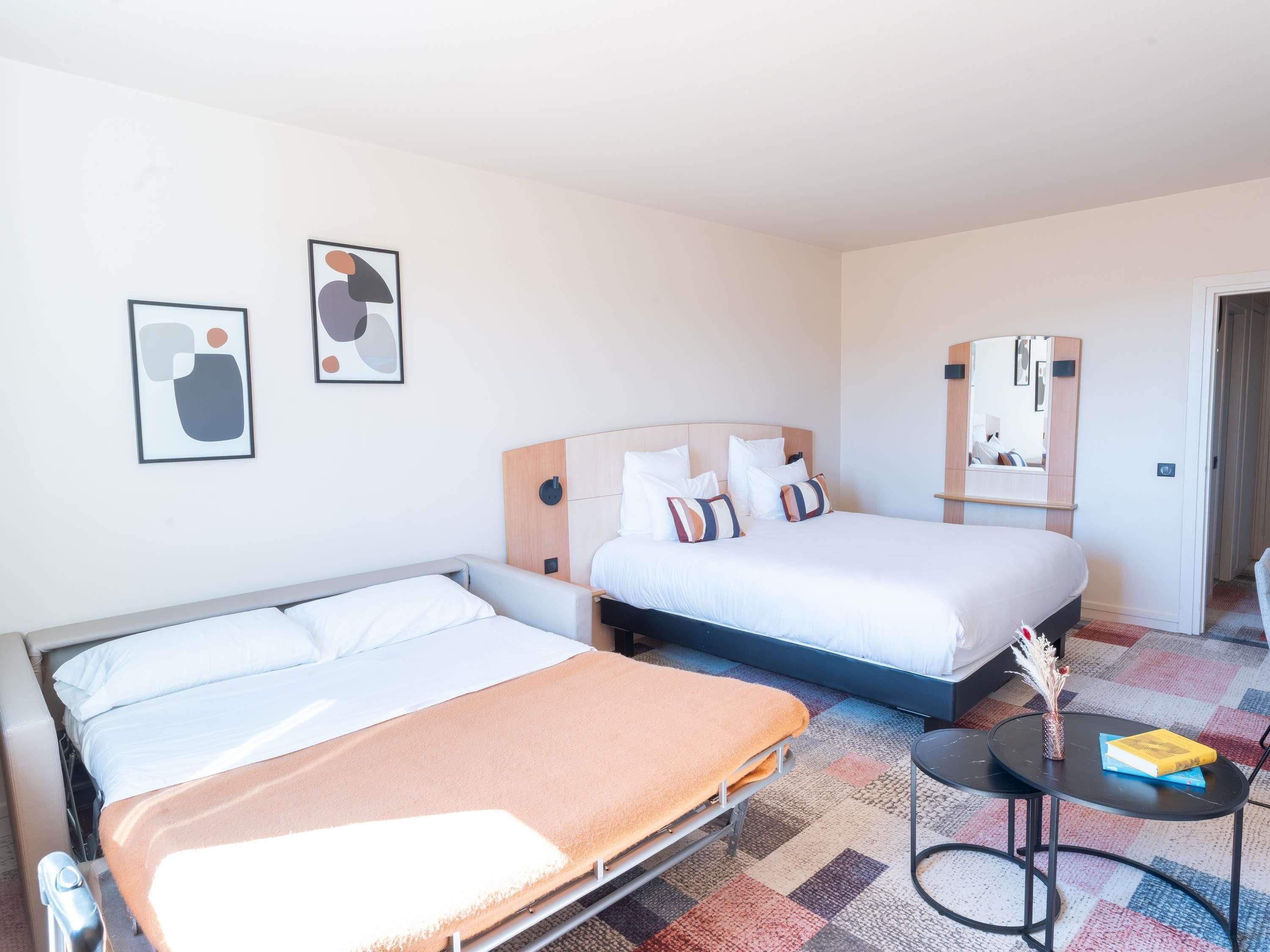 A hotel room with two beds, a small table, a mirror, and abstract art on the wall.