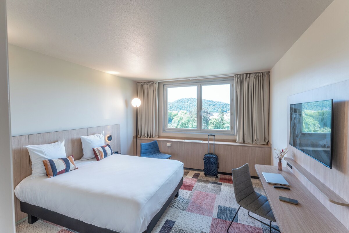 Habitación de hotel con una cama grande, un escritorio con una computadora portátil, un televisor y vistas a la montaña.