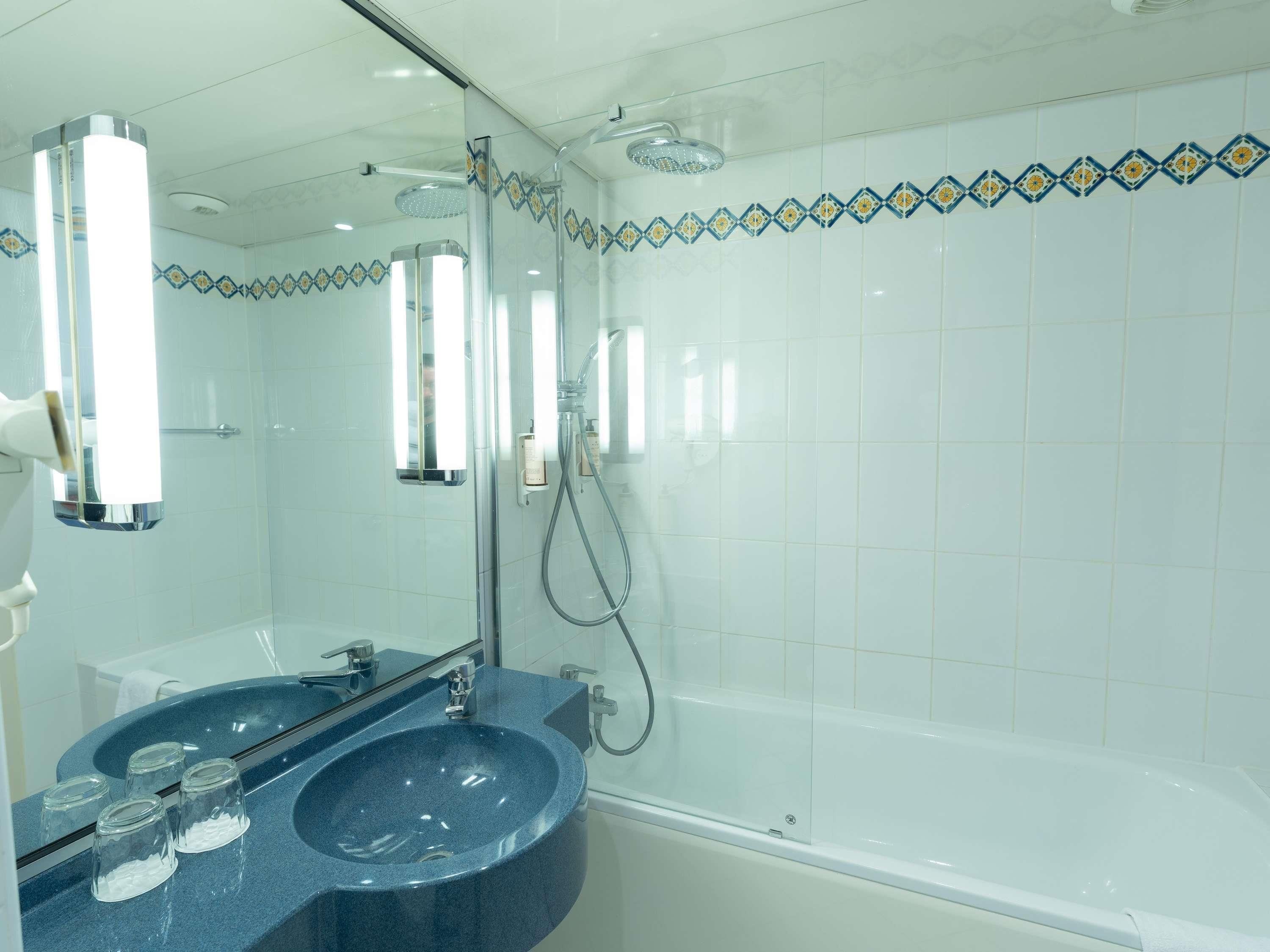 A modern bathroom with a dual sink vanity, a large mirror, a shower with a handheld showerhead, and decorative tile borders.
