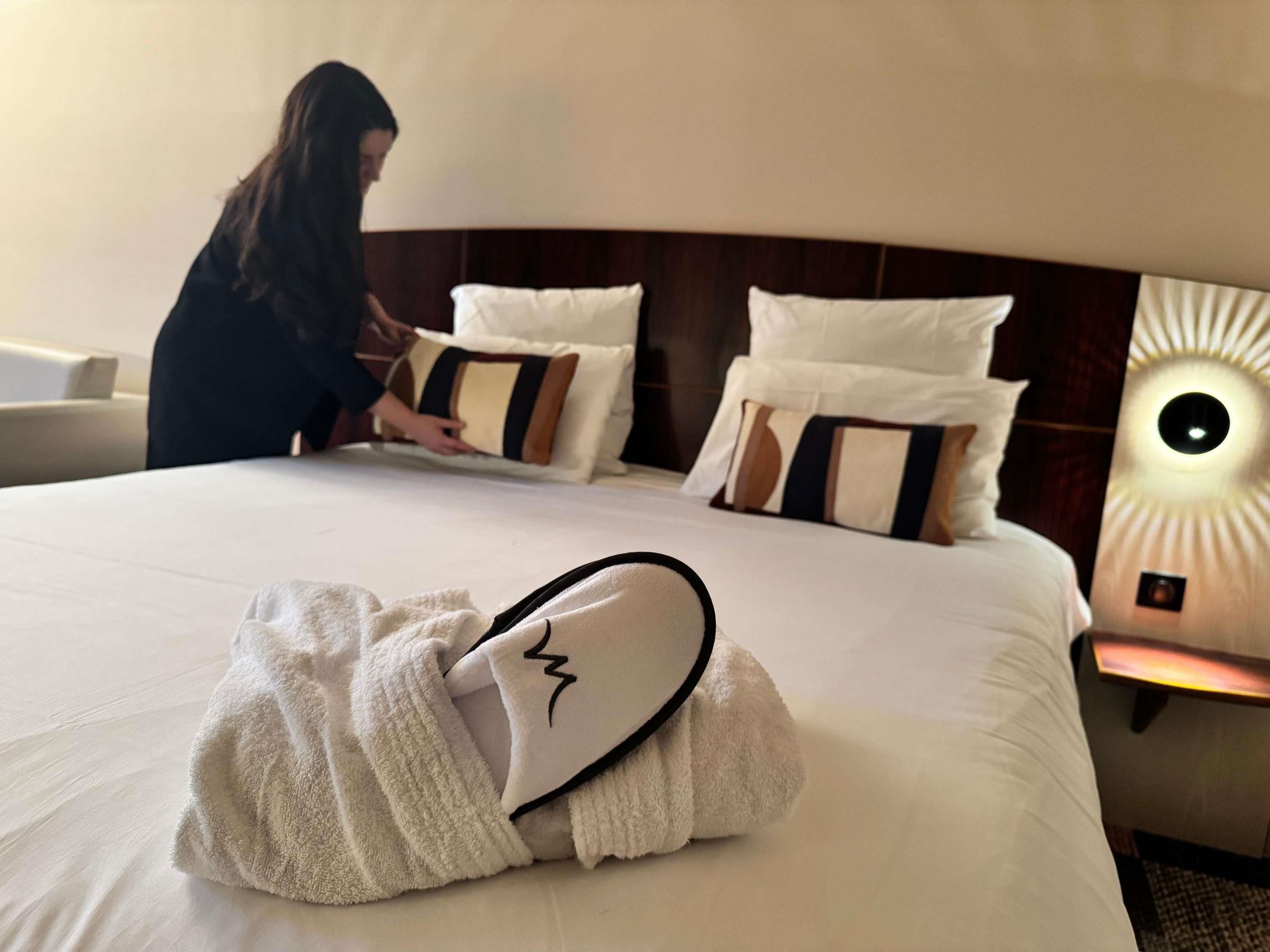 Une chambre d’hôtel avec un lit, des oreillers, une table de chevet avec une lampe, et une personne qui ajuste les oreillers.