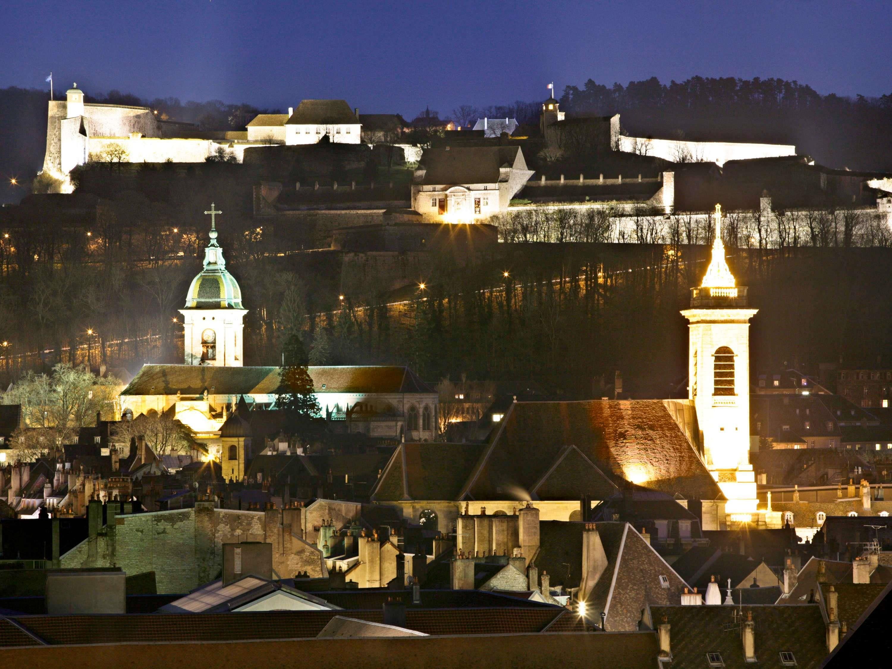 A historic town with a prominent castle on a hill, a church with a green dome, and a tall tower with a lit spire.