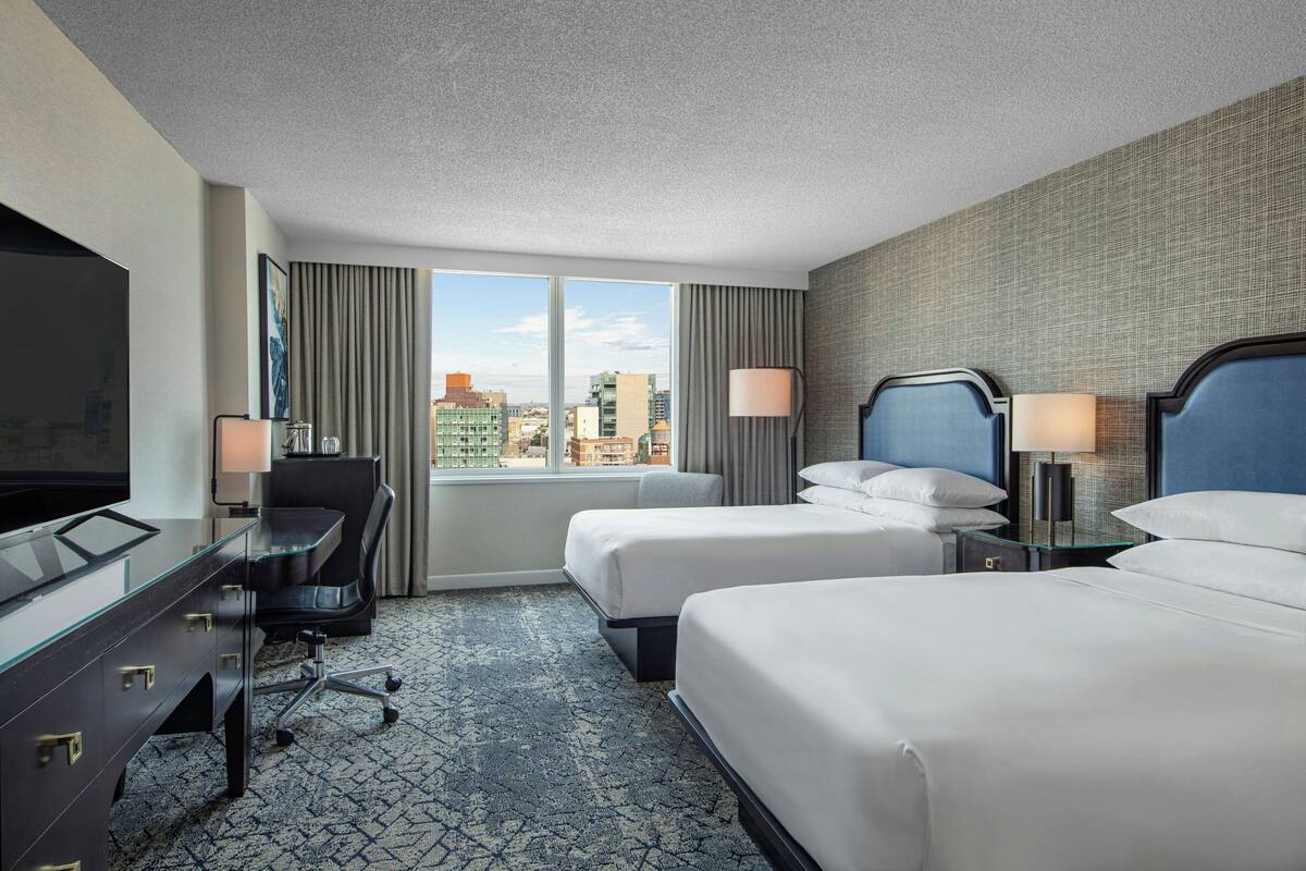 Habitación de hotel con dos camas, un escritorio con silla, televisión y vista a la ciudad.