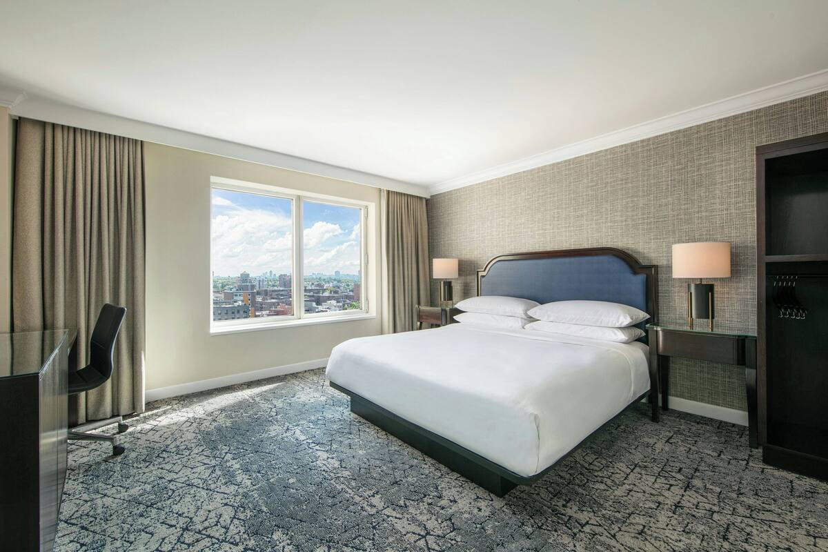 Habitación de hotel con una cama grande, dos mesitas de noche, un escritorio y una silla. Se ve una ventana con vista a la ciudad.