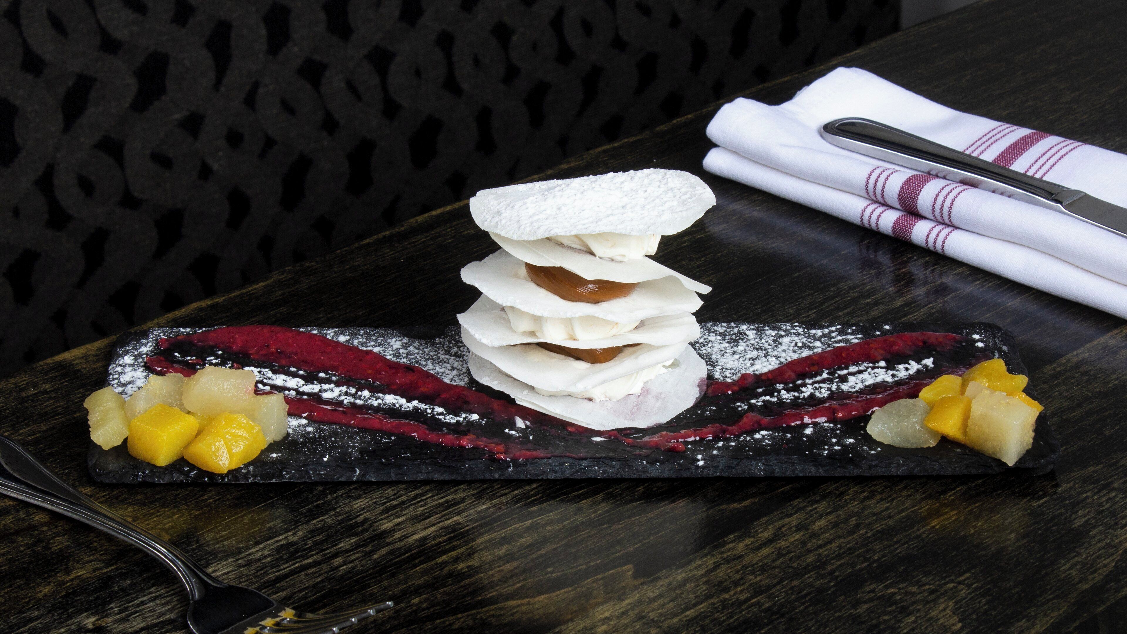 Postre sobre una losa de pizarra negra, con azúcar glas, un cuchillo y una servilleta sobre una mesa de madera oscura.