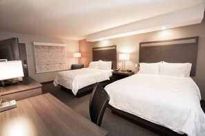 A hotel room with two beds, a desk, a chair, a TV, and a lamp.
