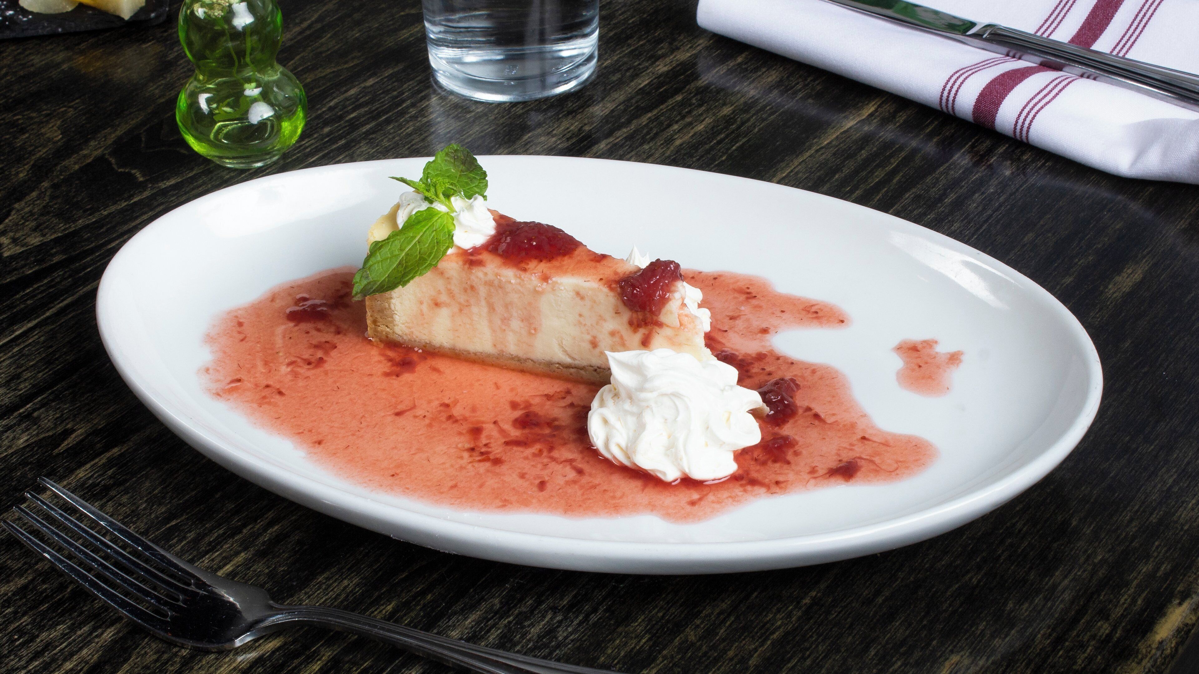 Une assiette à dessert avec une part de gâteau au fromage, de la crème fouettée et une sauce aux fruits rouges, le tout posé sur une table en bois foncé.