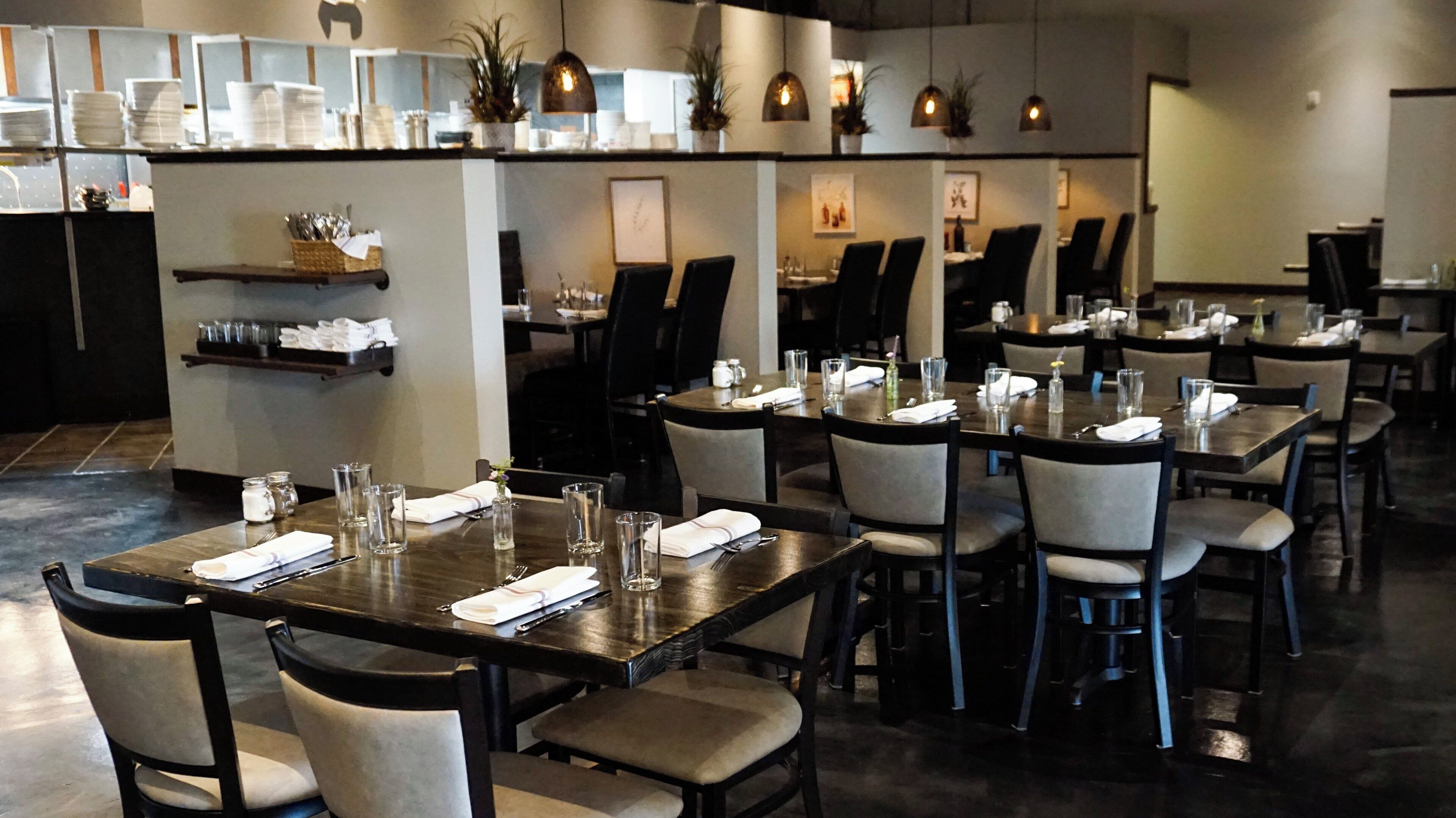 A restaurant with multiple tables set for dining, chairs, and a counter area with stacked plates and glasses.