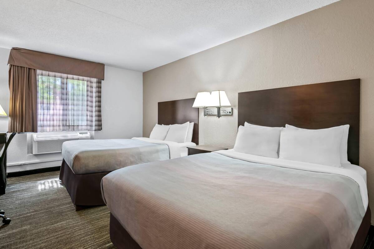 A hotel room with two beds, a desk, a chair, a window with curtains, and a wall-mounted lamp.