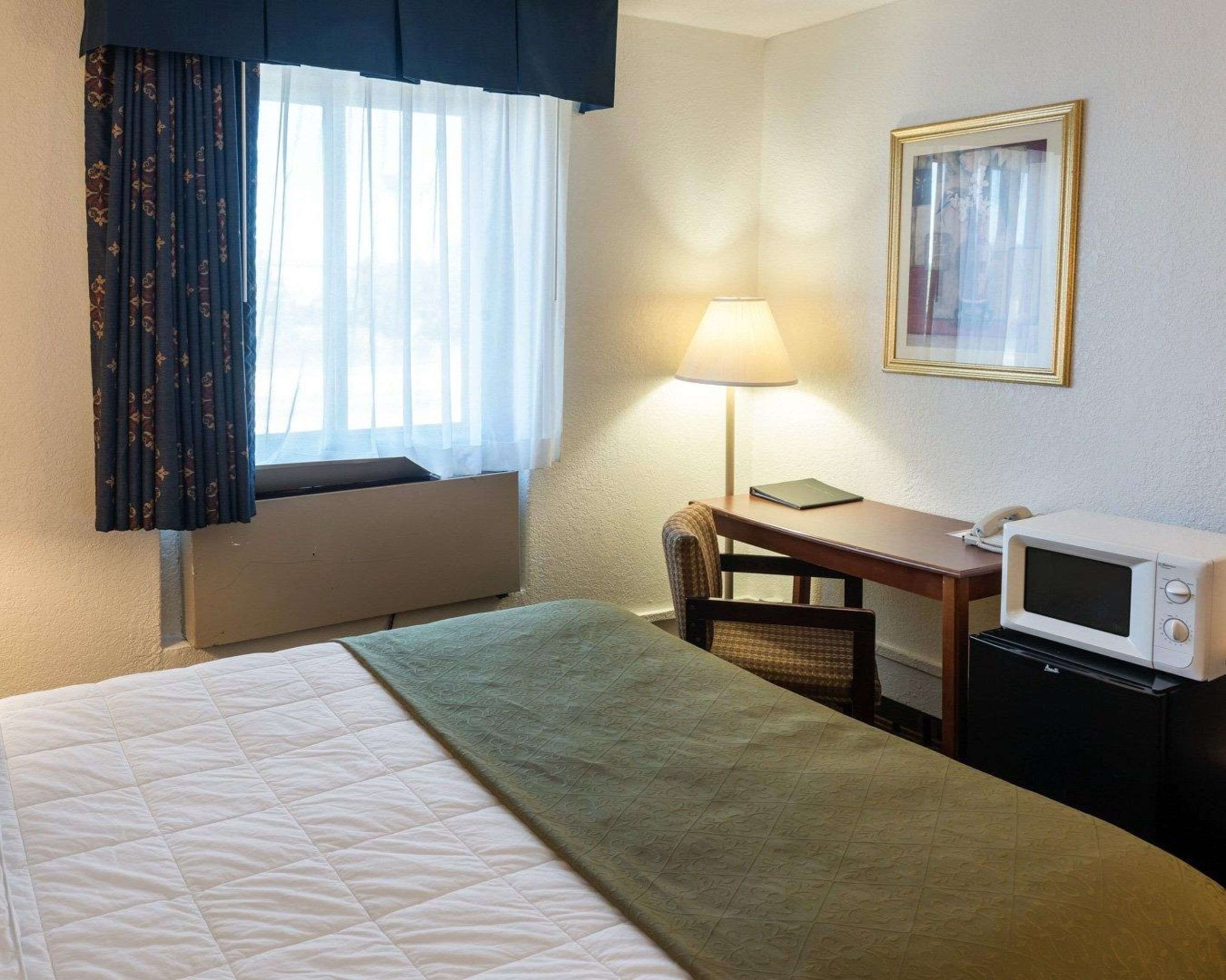 Ein Hotelzimmer mit einem Bett, einem Schreibtisch, einer Lampe, einer Mikrowelle und einem gerahmten Bild an der Wand.