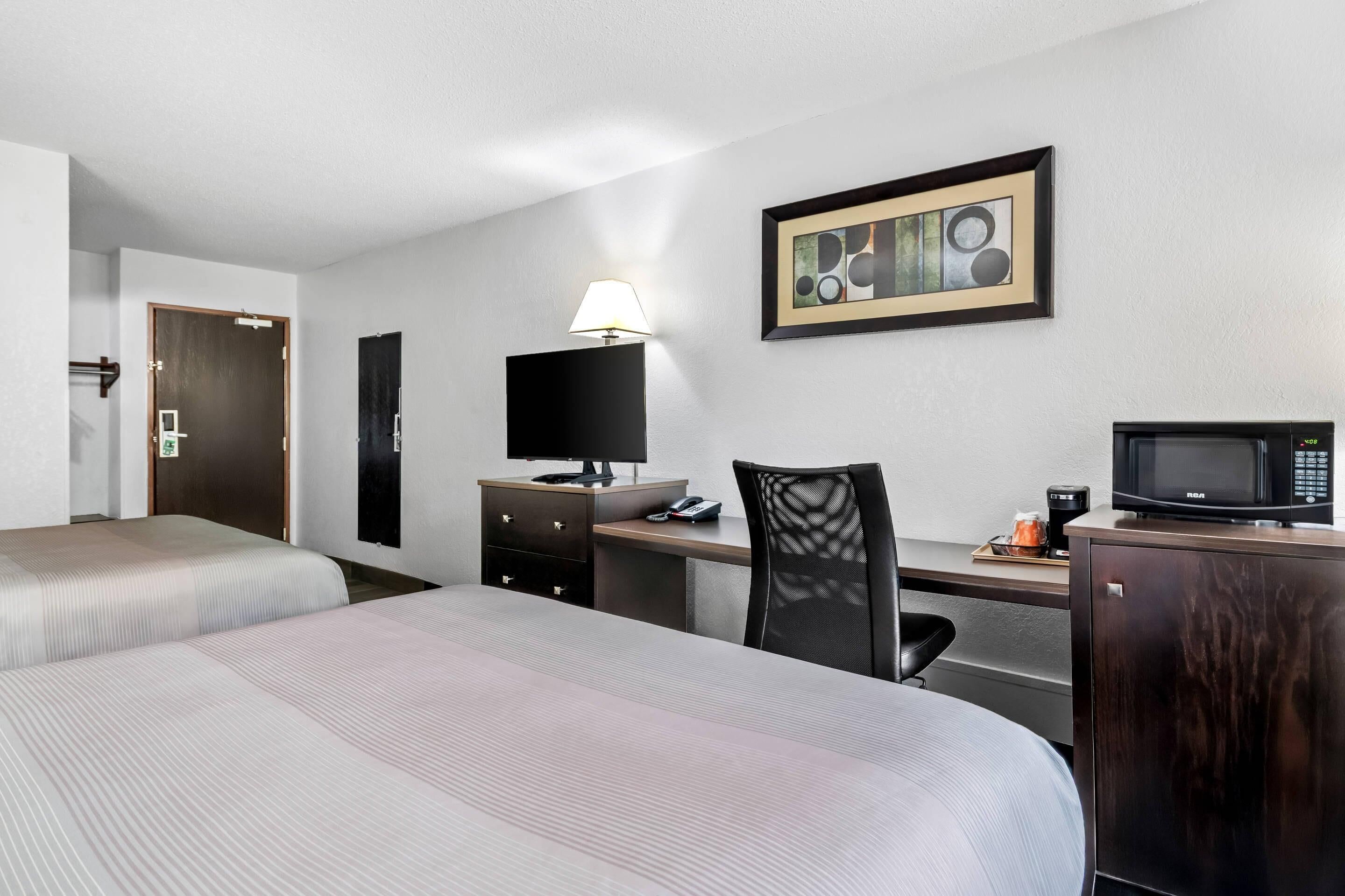 A hotel room with two beds, a desk with a computer, a chair, a desk lamp, a microwave, and a framed picture on the wall.