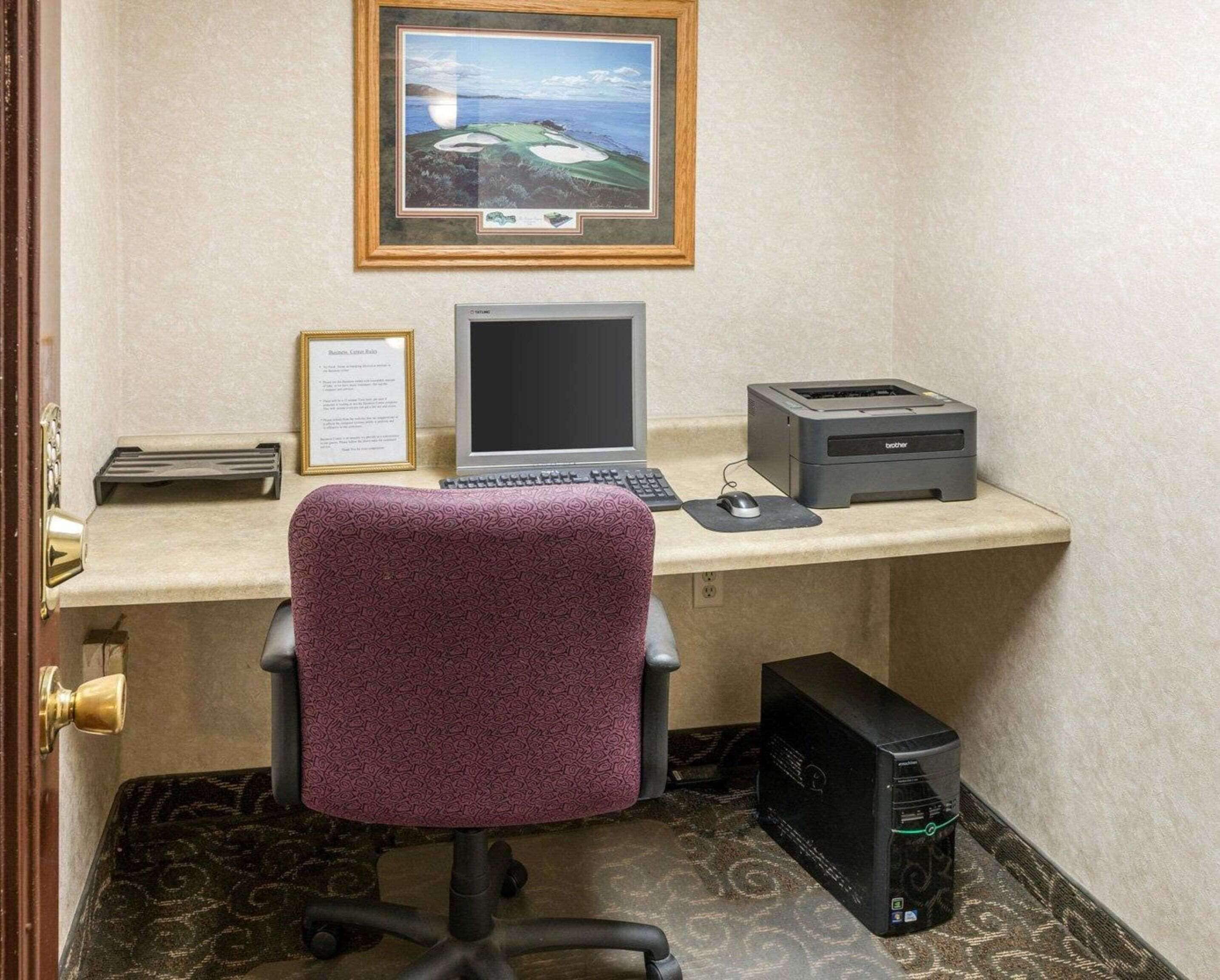 A hotel room with a desk, a laptop, a printer, a framed certificate, and a desktop computer.