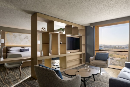 A modern hotel room with a large TV, comfortable seating, a bookshelf, and a city view.