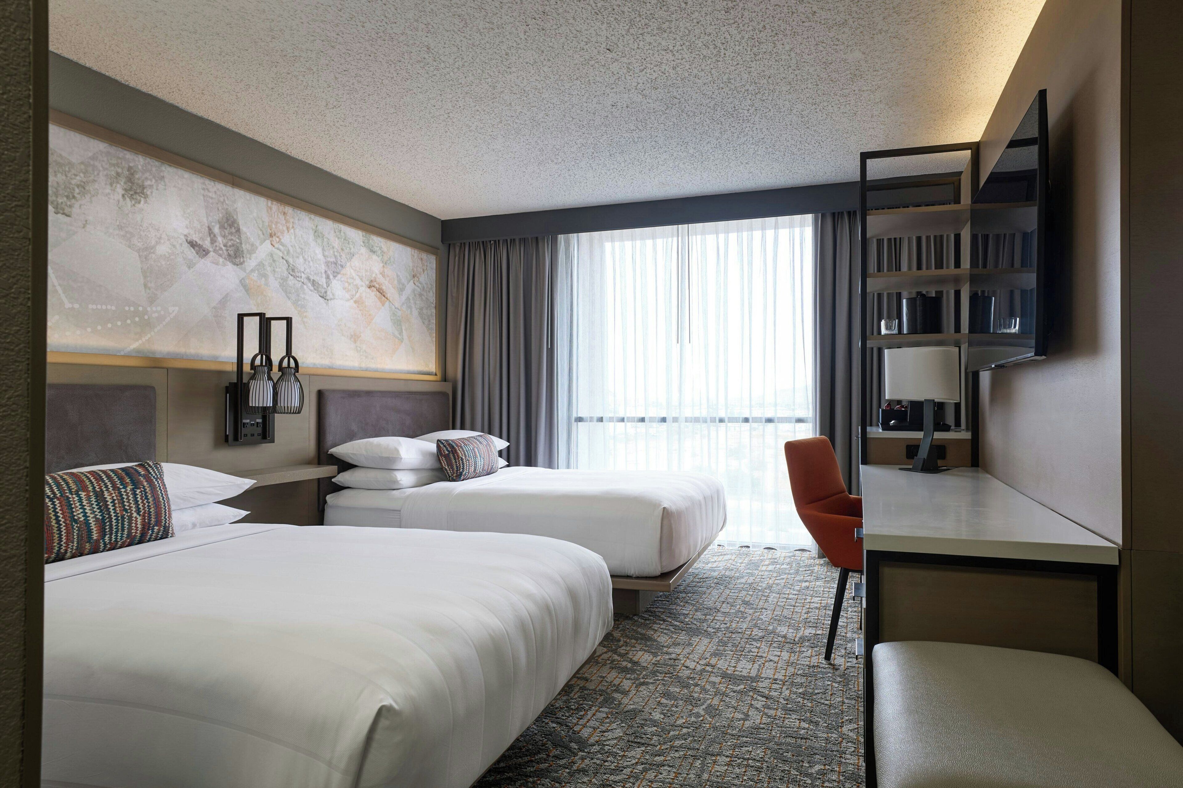 A hotel room with two beds, a desk, a chair, a TV, and a large window with curtains.