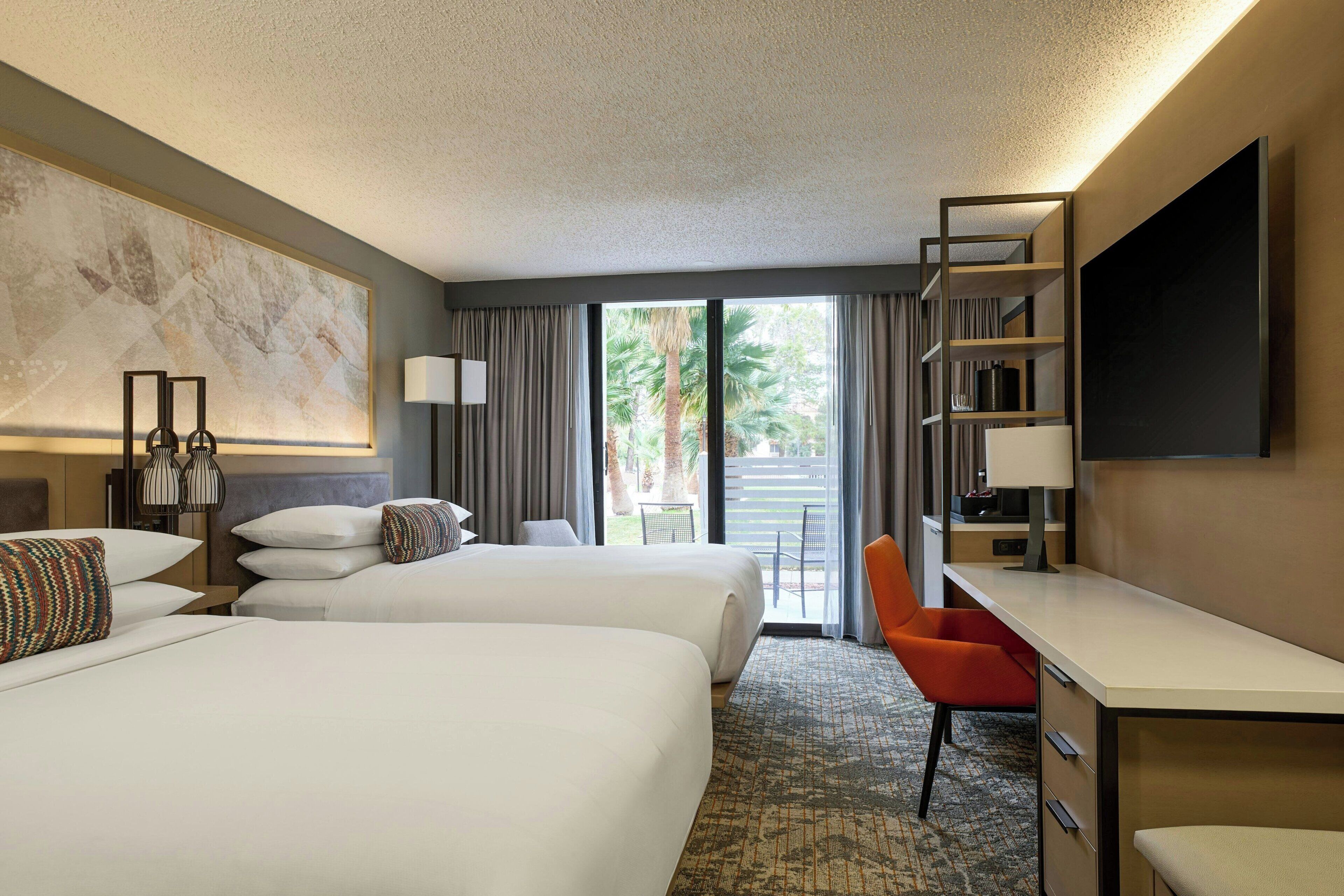 A hotel room with two beds, a desk, a chair, a TV, and a balcony with a view of palm trees.
