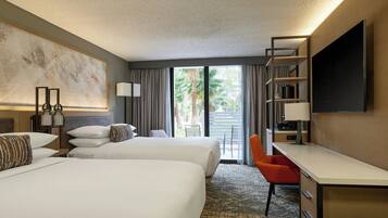 A hotel room with two beds, a desk, a chair, a TV, and a balcony with a view of palm trees.