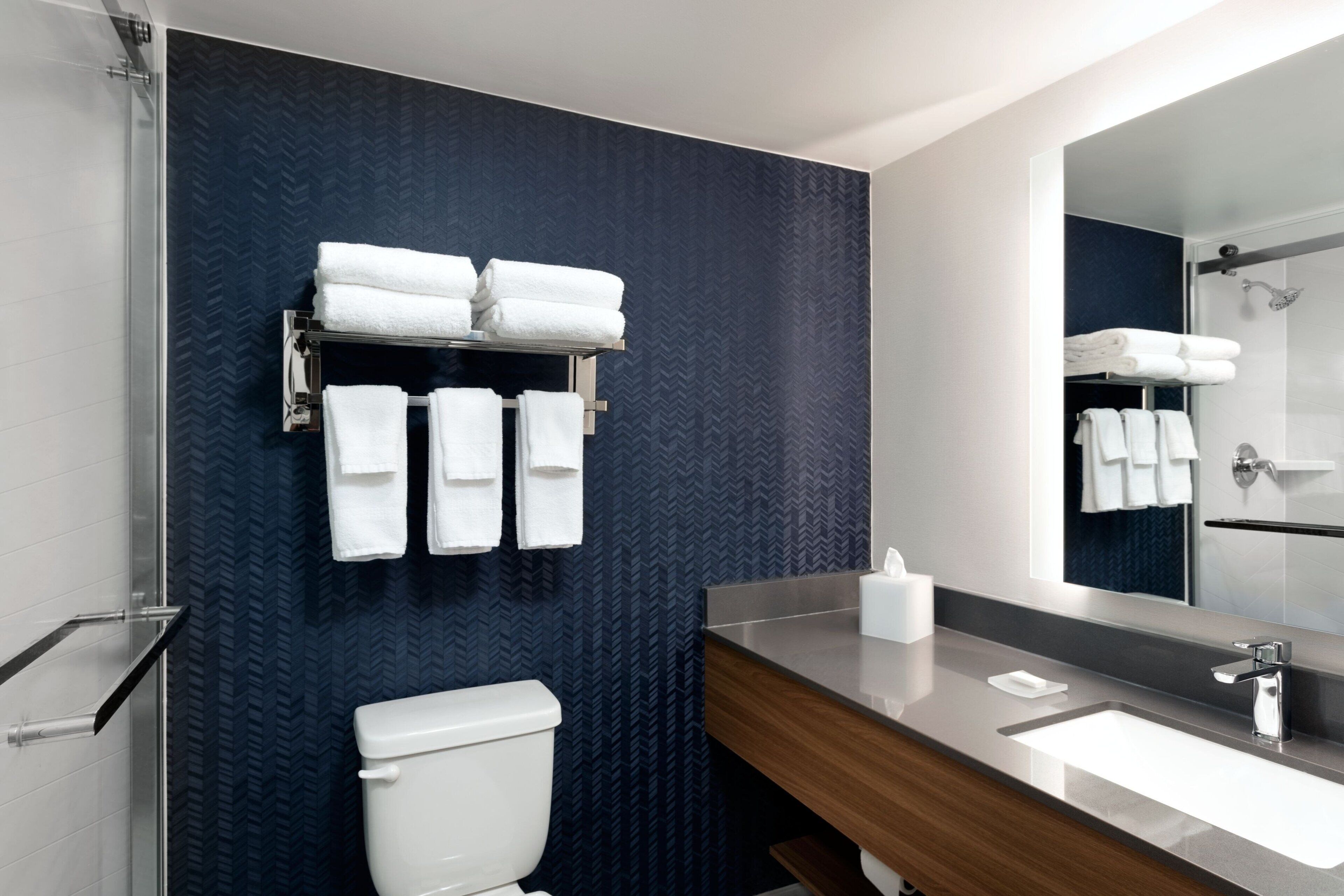 Ein modernes Badezimmer mit einem großen Spiegel, einer weißen Toilette, einem Waschbecken mit Holzauskleidung und einem Handtuchhalter mit ordentlich gefalteten Handtüchern.